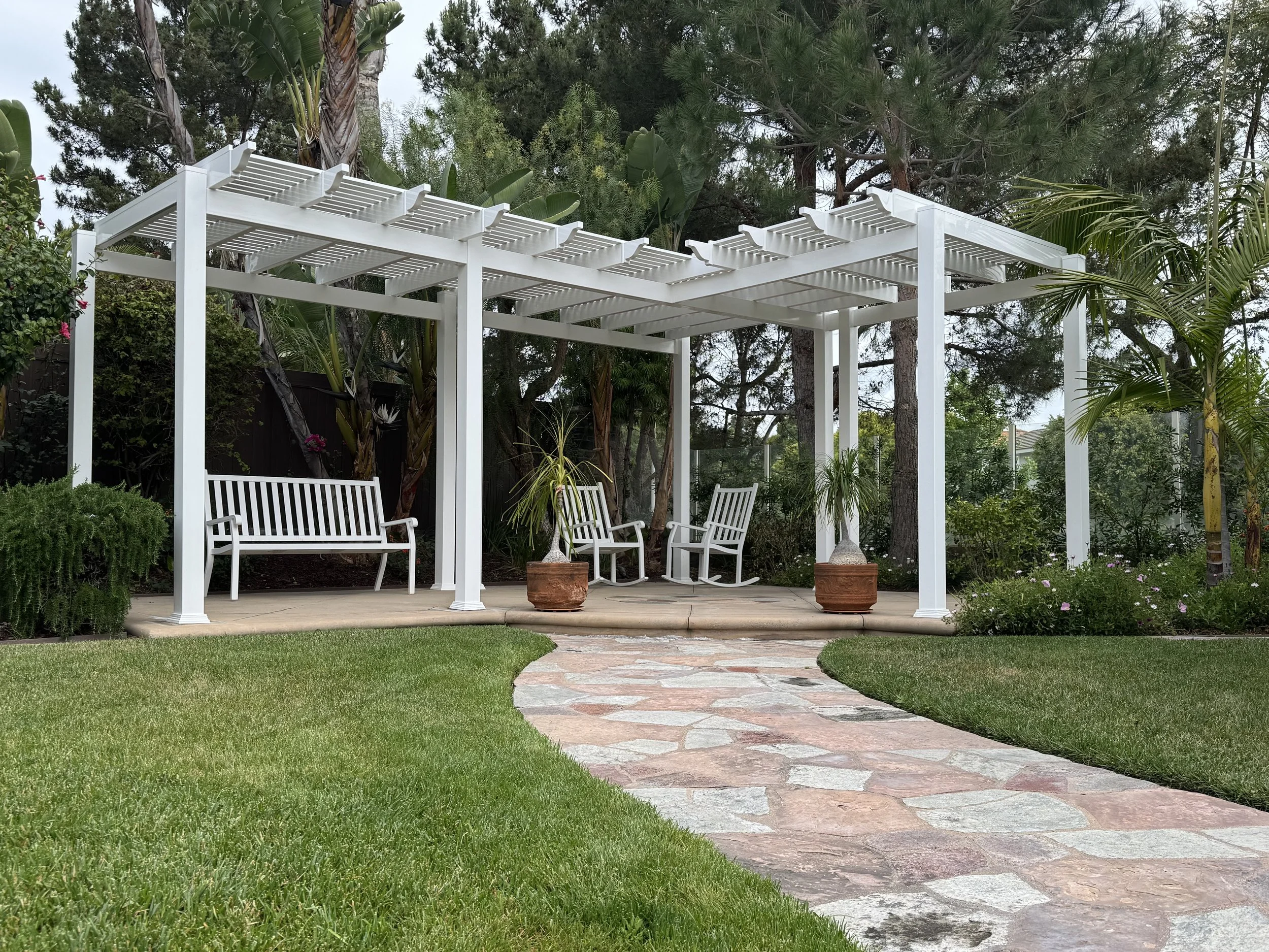 A white pergola with three chairs and two potted plants in a garden with grass and a stone pathway.