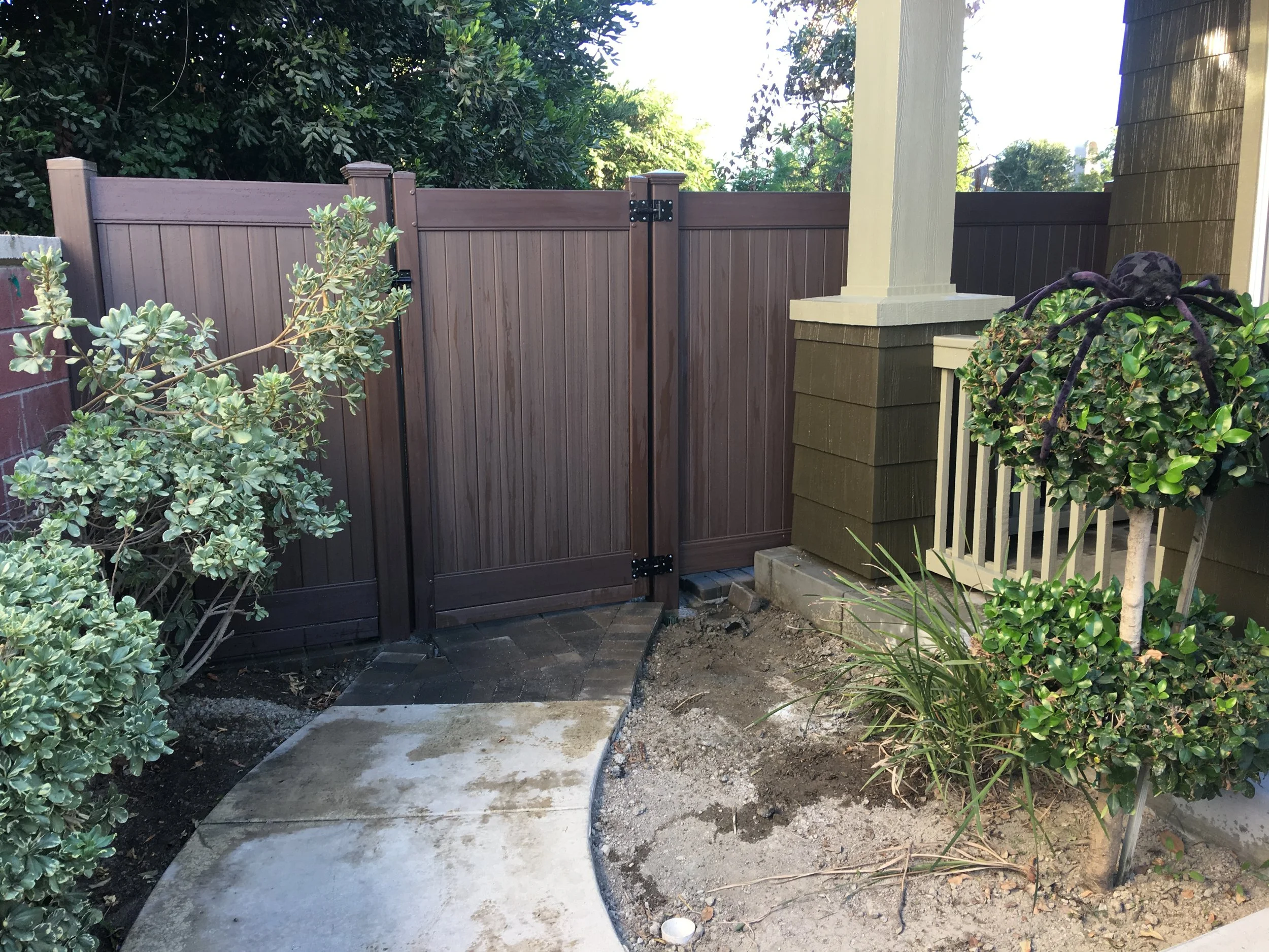 La Habra backyard corner with brown vinyl privacy fence, curved concrete pathway and manicured landscape featuring green bushes and purple flower pot under porch railing, highlighting stylish vinyl fences and gates in Orange County.