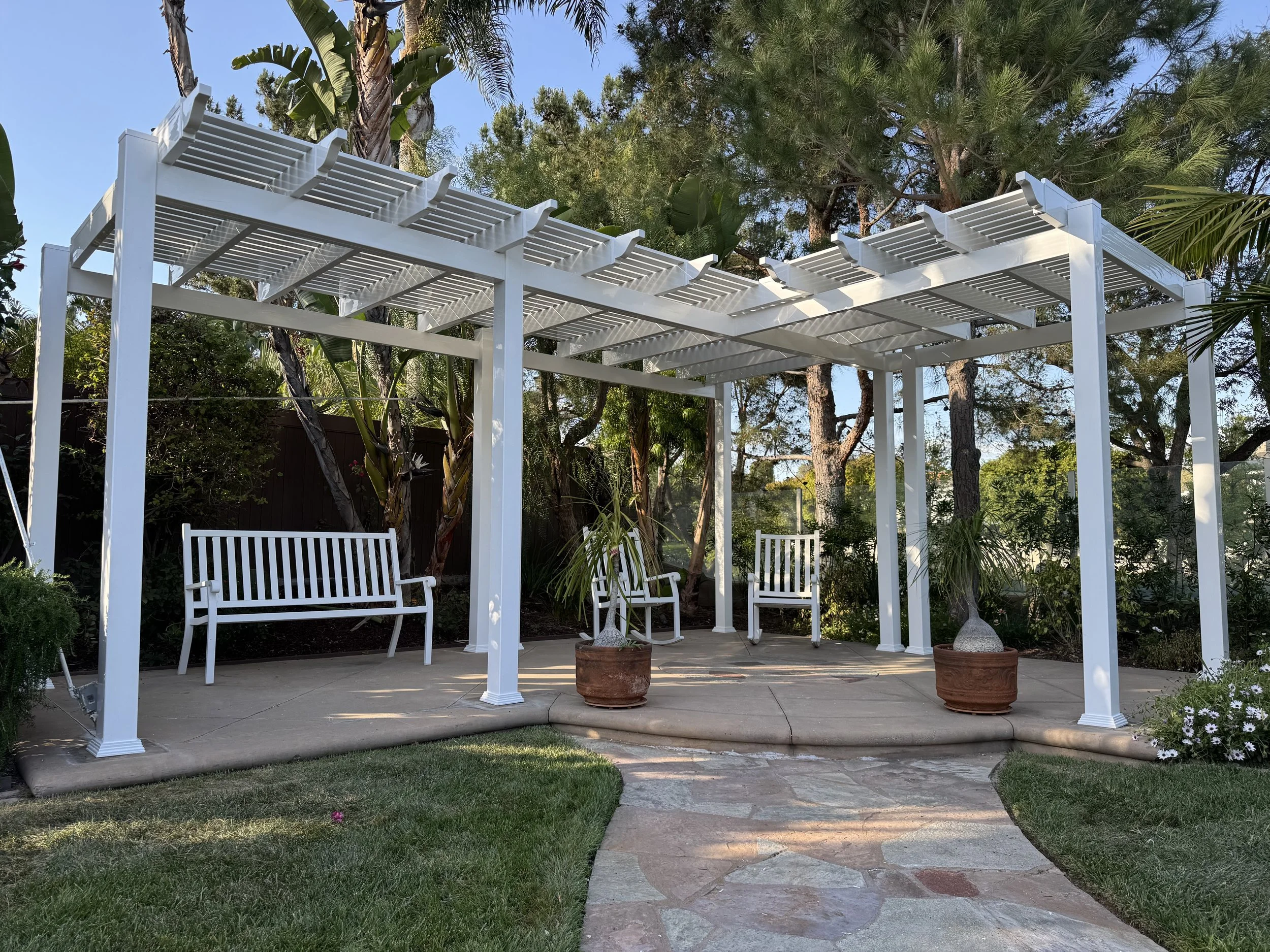 Yorba Linda white vinyl pergola with benches and potted plants on a stone patio in a lush garden with trees and shrubs, highlighting elegant vinyl patio covers and fencing in Orange County.