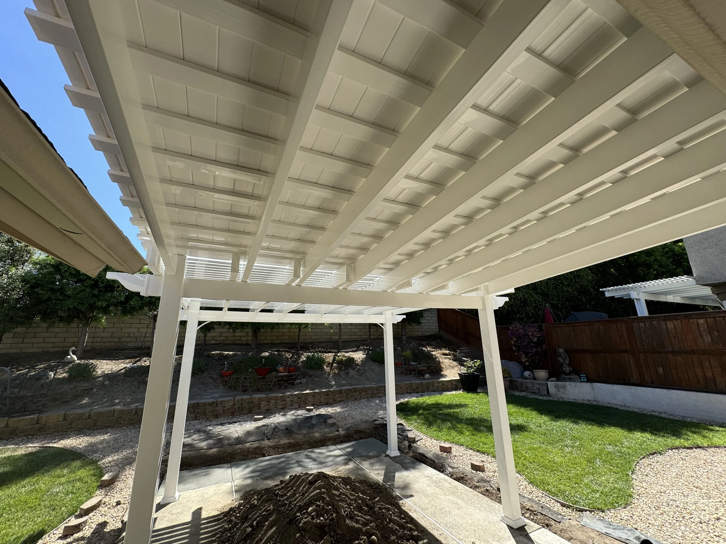 View of a backyard patio area with a white pergola, green grass, a brick wall, and trees in the background.