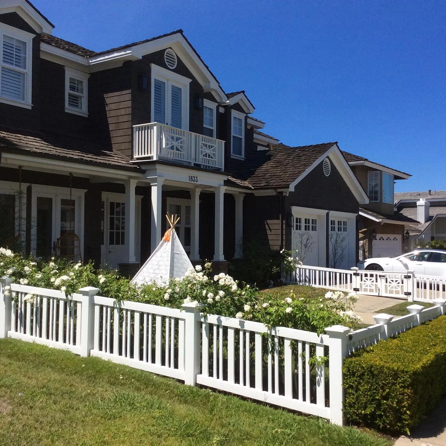 Aliso Viejo two-story modern house with dark exterior siding, white trim, and a small front porch with white columns. White picket fence surrounds a garden with white flowers and a small teepee, ideal for showcasing custom vinyl gates and fences.