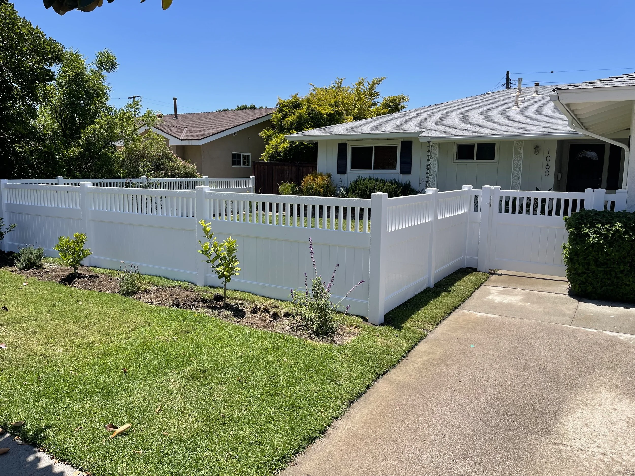 Garden Grove front yard with lush green plants and shrubs surrounded by white vinyl privacy fence and concrete driveway leading to a gray-roofed house with blue shutters, illustrating premium vinyl fences for secure Orange County homes.