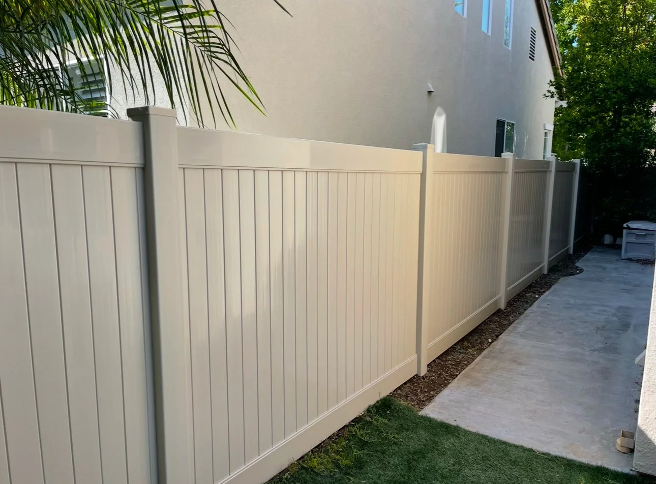 Tall white vinyl privacy fence runs along a concrete sidewalk next to a beige house with multiple windows, with trees and greenery on the side, demonstrating durable vinyl fencing in Orange Ca.