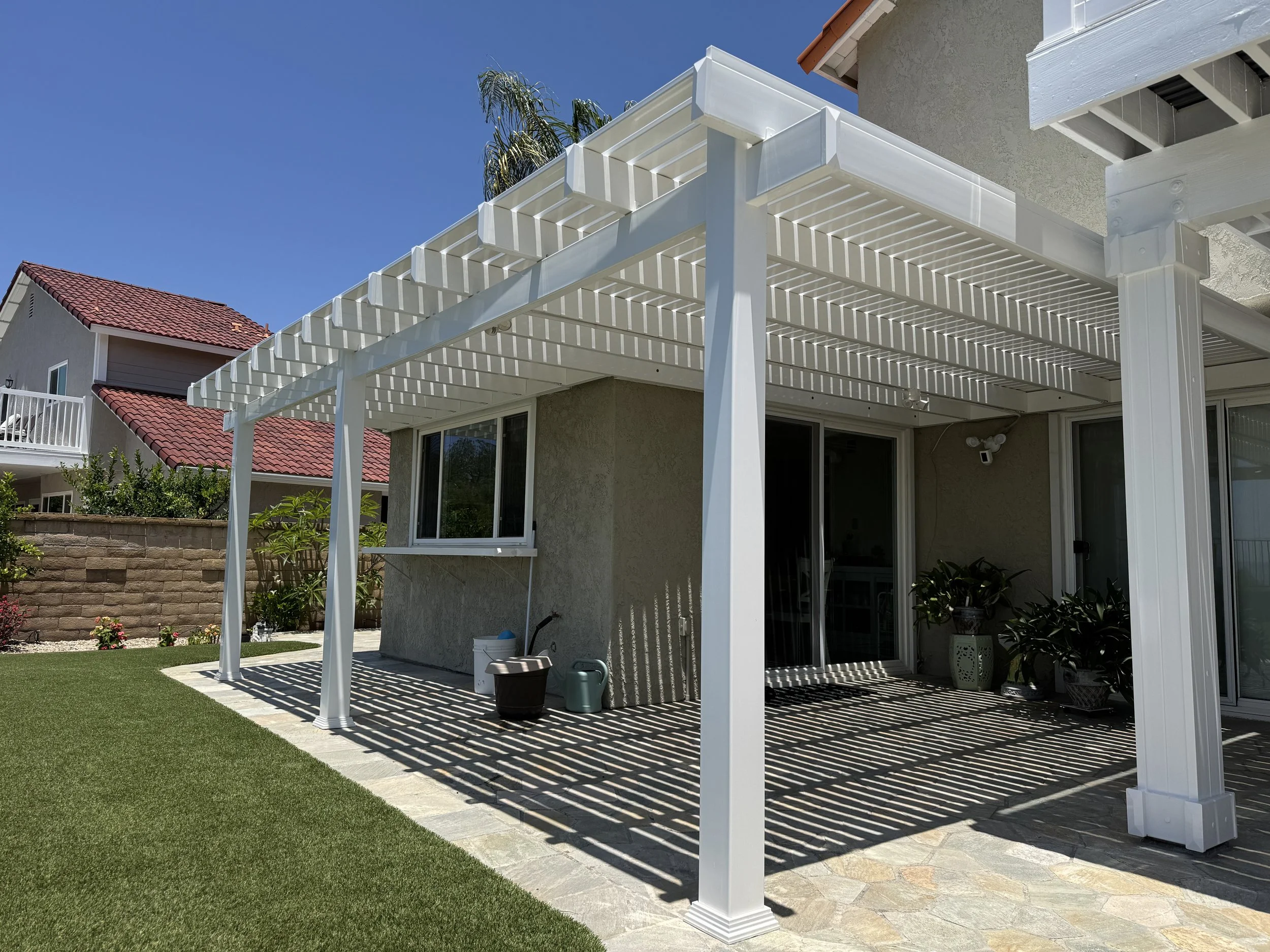 Backyard patio with a white wooden pergola casting striped shadows, adjacent to a house with sliding glass doors, outdoor plants, and gardening supplies.