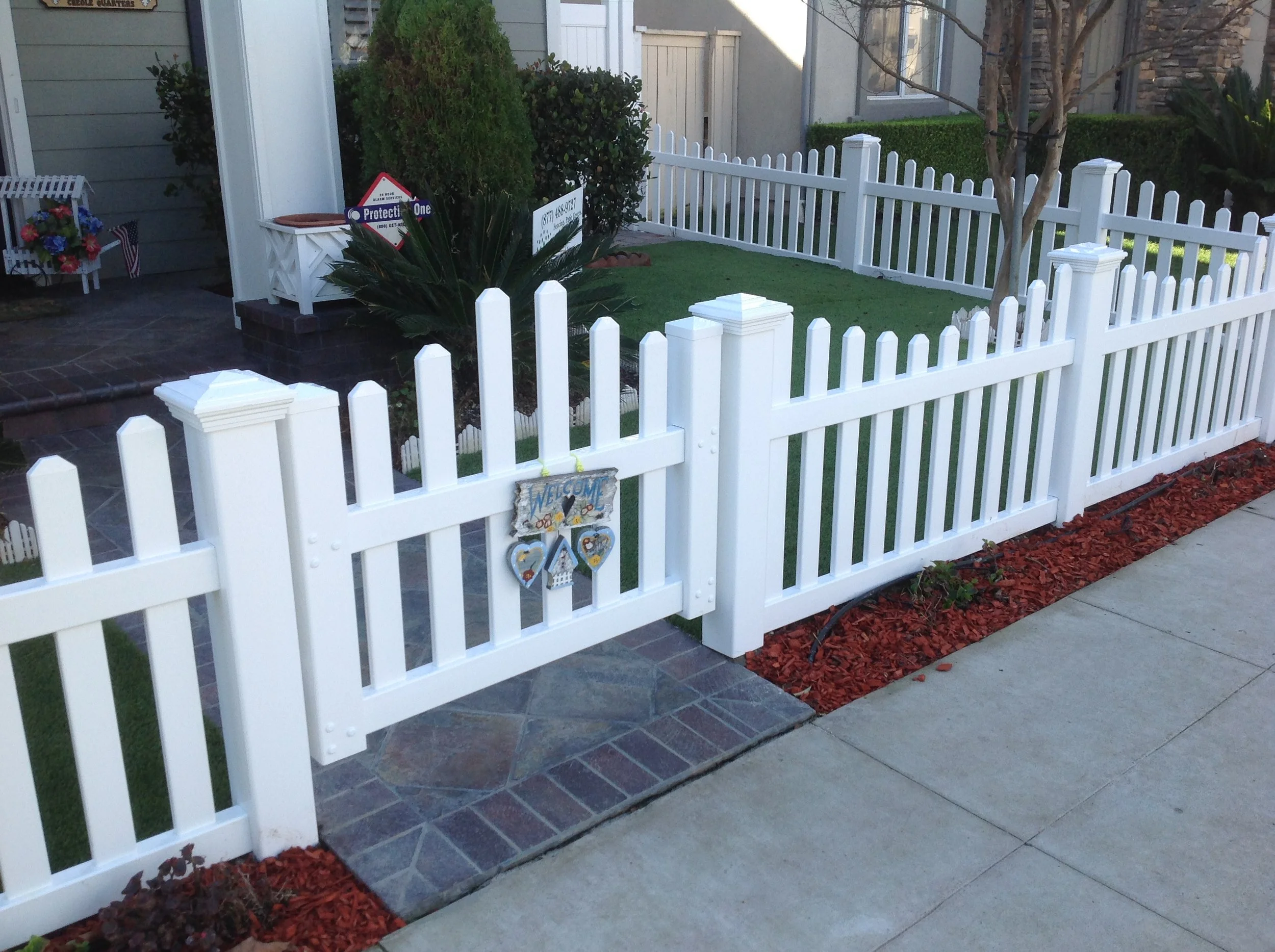Tustin white vinyl picket fence with arched pedestrian gate and decorative posts, stone paver step and red mulch bordering a neat front yard.