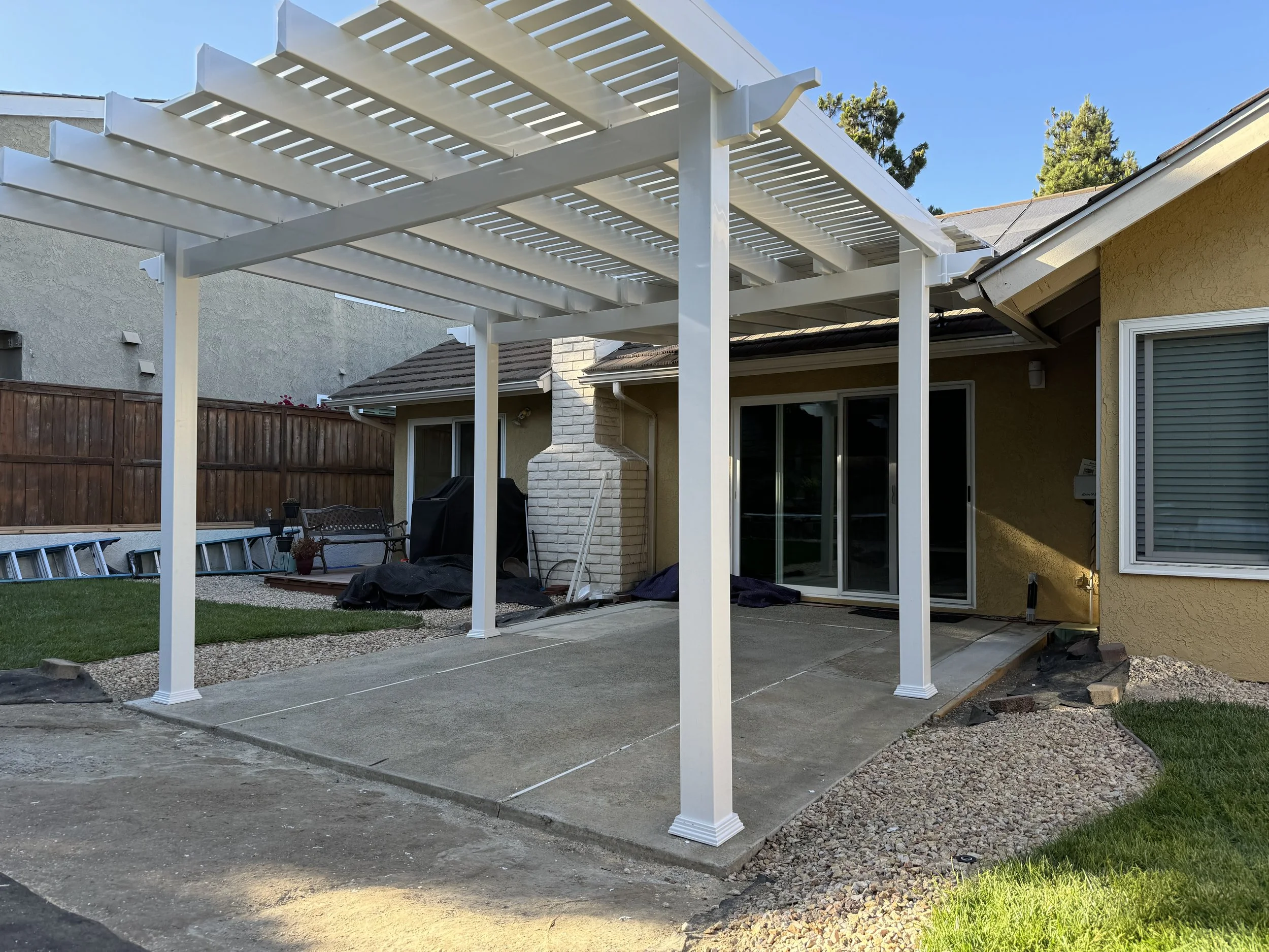 Irvine backyard patio with a white vinyl pergola and concrete floor, featuring a sliding glass door, window, and grill covered with a tarp, with a grassy area and neighboring houses, showing how vinyl patio covers enhance outdoor living in Orange Cou