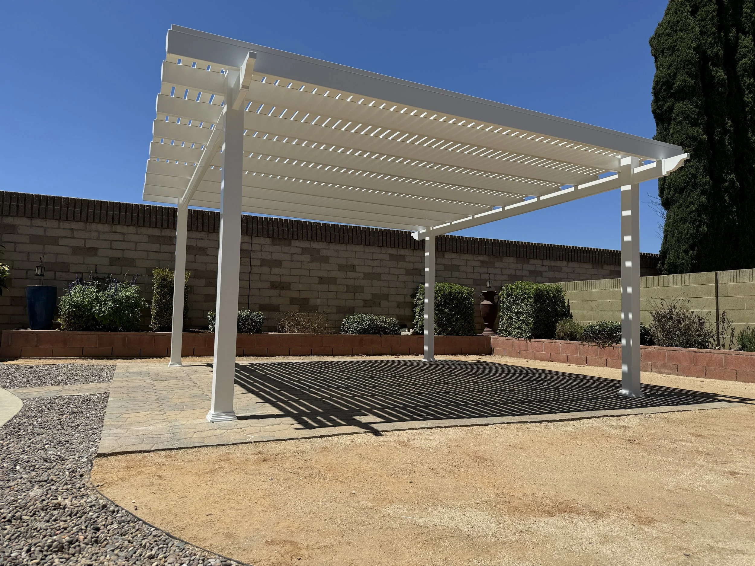 Newport Beach white vinyl pergola with horizontal slats over a brick and paving stone patio in a backyard, with a brick wall, plants, and trees under a clear blue sky, demonstrating stylish vinyl patio covers for coastal Orange County homes.
