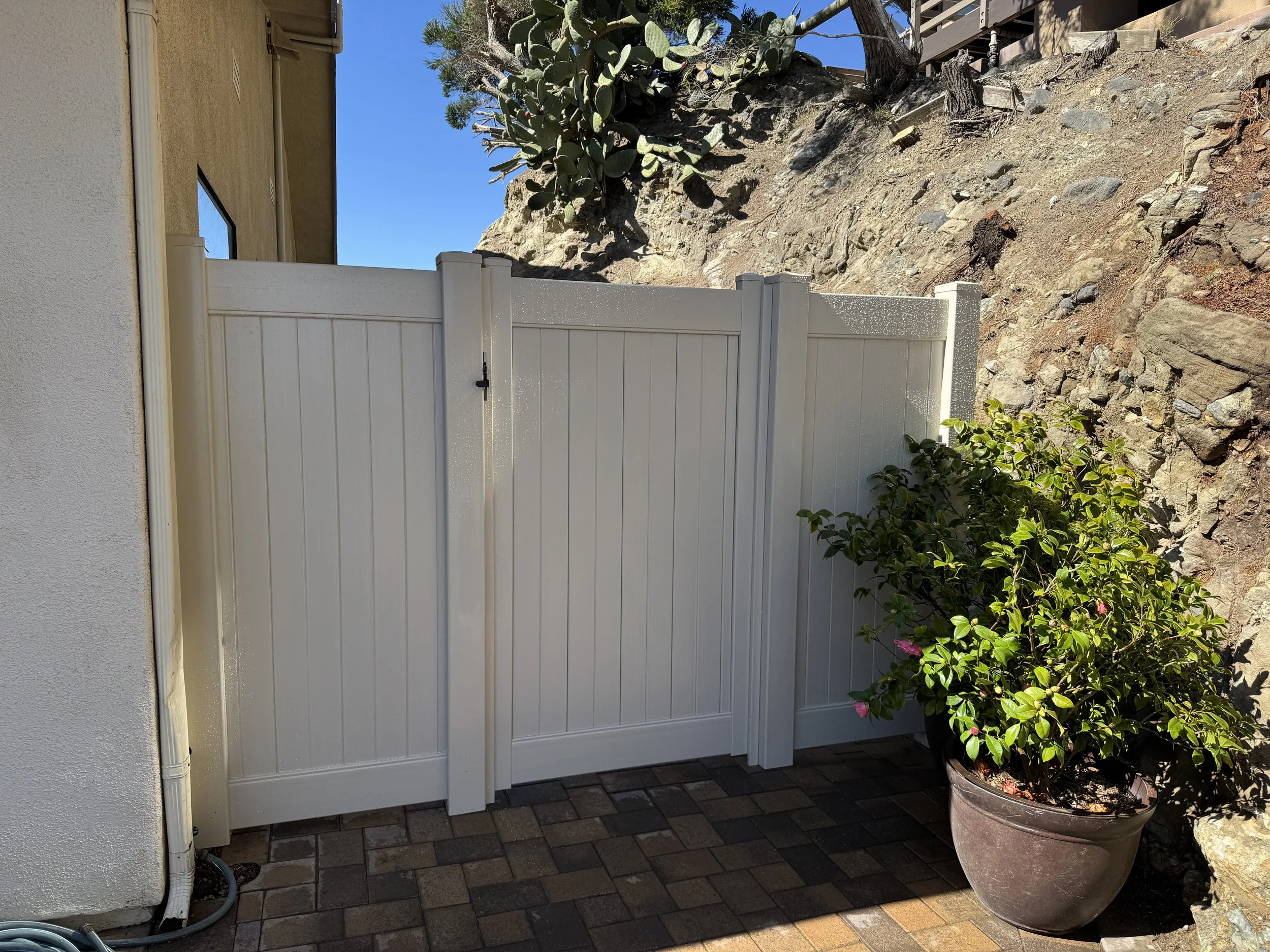 San Clemente white vinyl privacy fence and matching gate installed beside a stucco wall and rock hillside, with paver walkway and potted plant, durable low-maintenance fencing solution for coastal Orange County homes.