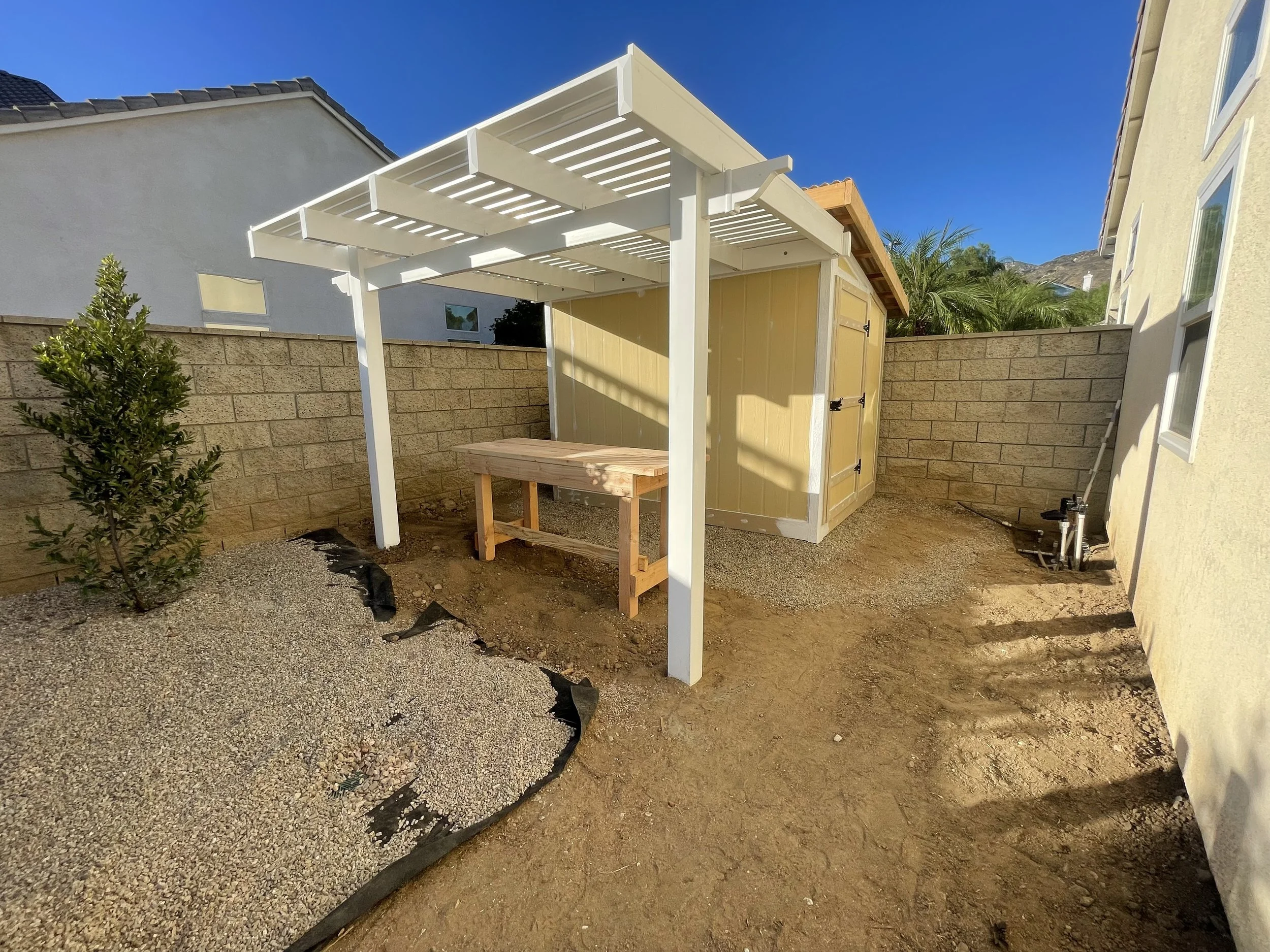 Backyard with a small yellow shed, a white pergola, and a wooden work table, with a brick wall and some plants in the background.