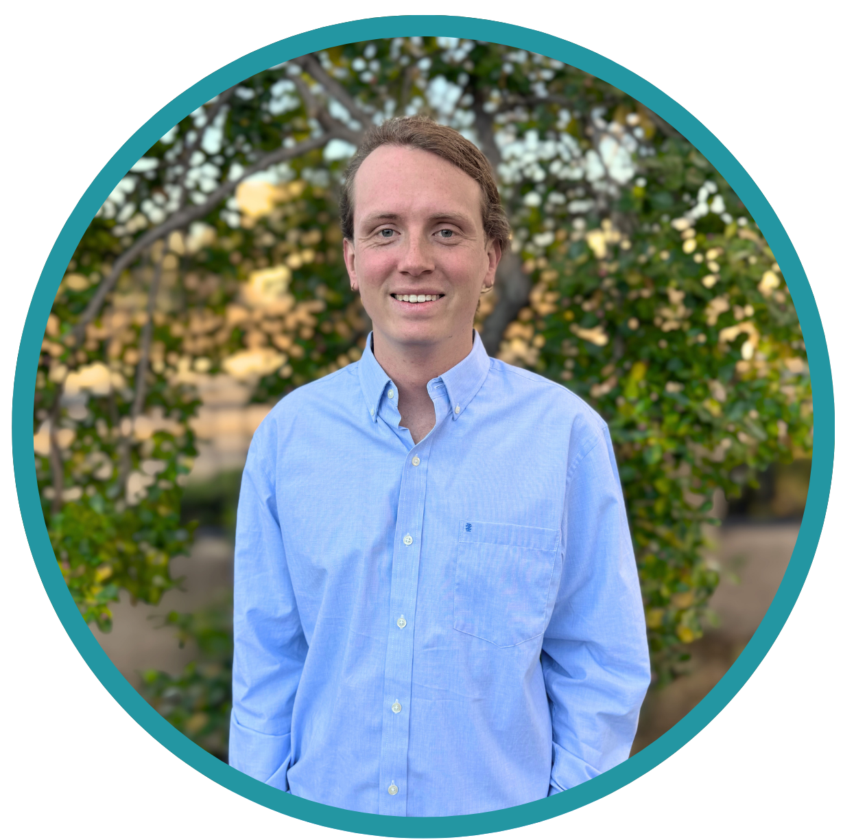 A young man with light brown hair, wearing a light blue button-up shirt, standing outdoors in front of a leafy green tree during daytime.