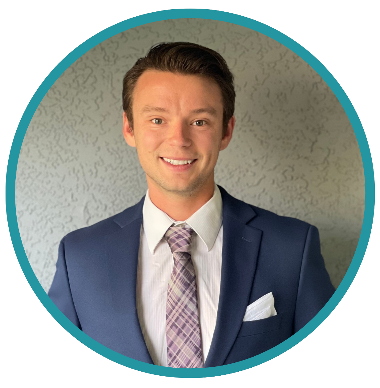 A smiling man in a navy suit, white shirt, and patterned tie, with a white pocket square, standing against a textured gray wall.