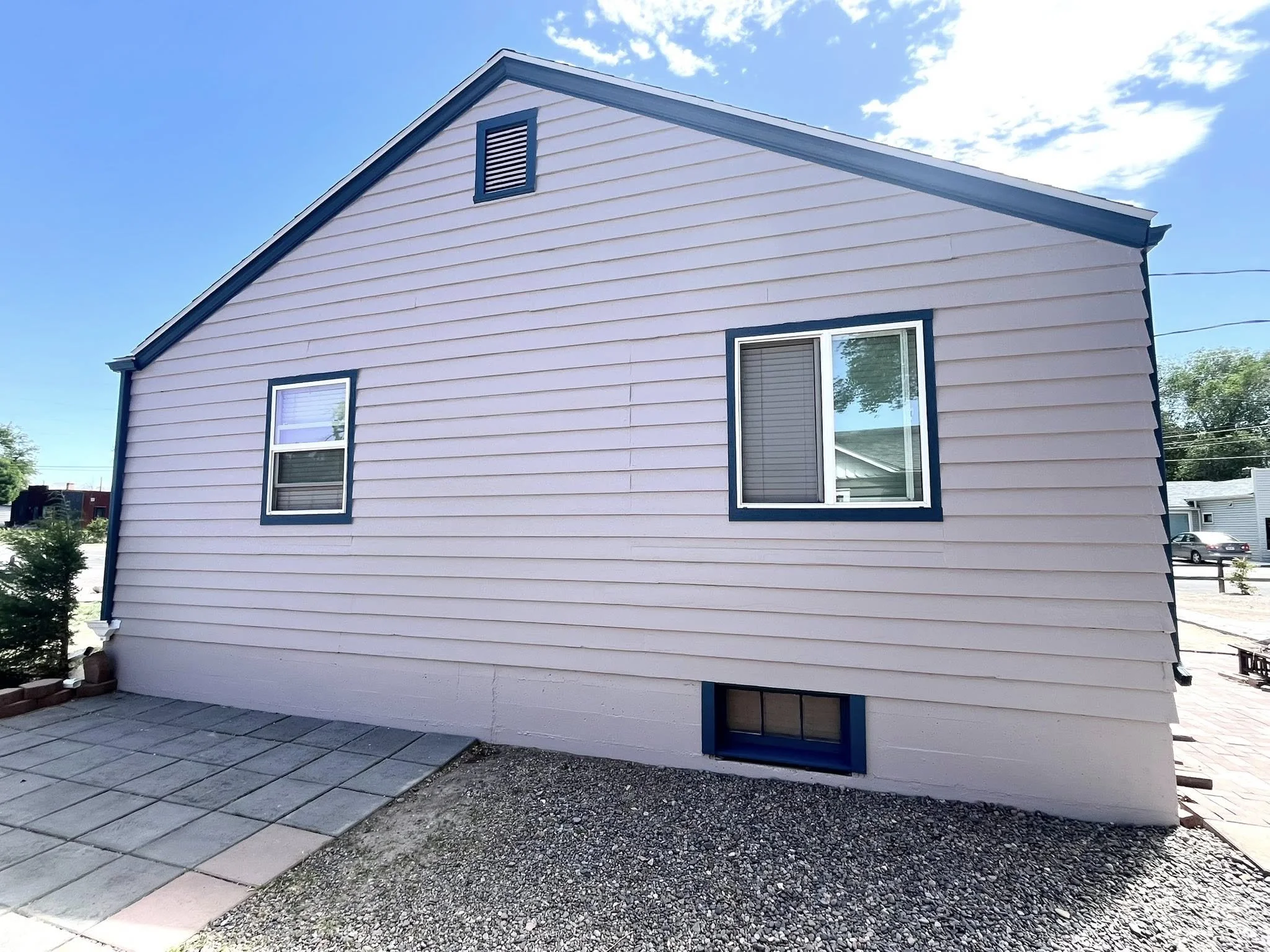 Side view of a beige house with blue trim, two windows on the upper floor, and a small basement window, with a paved area and gravel in the front yard, under a partly cloudy sky.