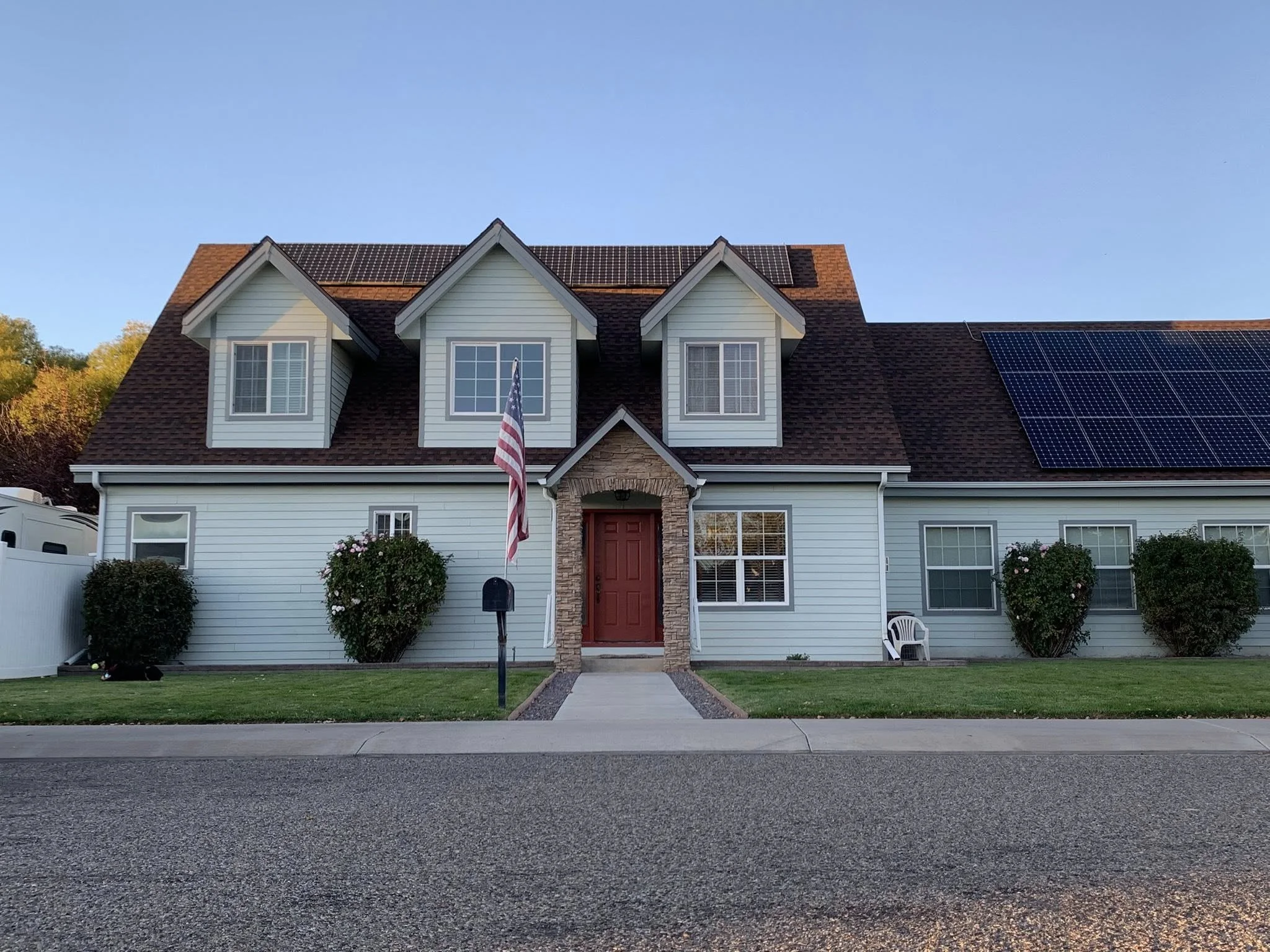 A two-story house with white siding, a brown shingled roof with solar panels, three dormer windows, and a red front door surrounded by a stone arch. There is an American flag near the entrance, three bushes in front, and a white lawn chair on the rig