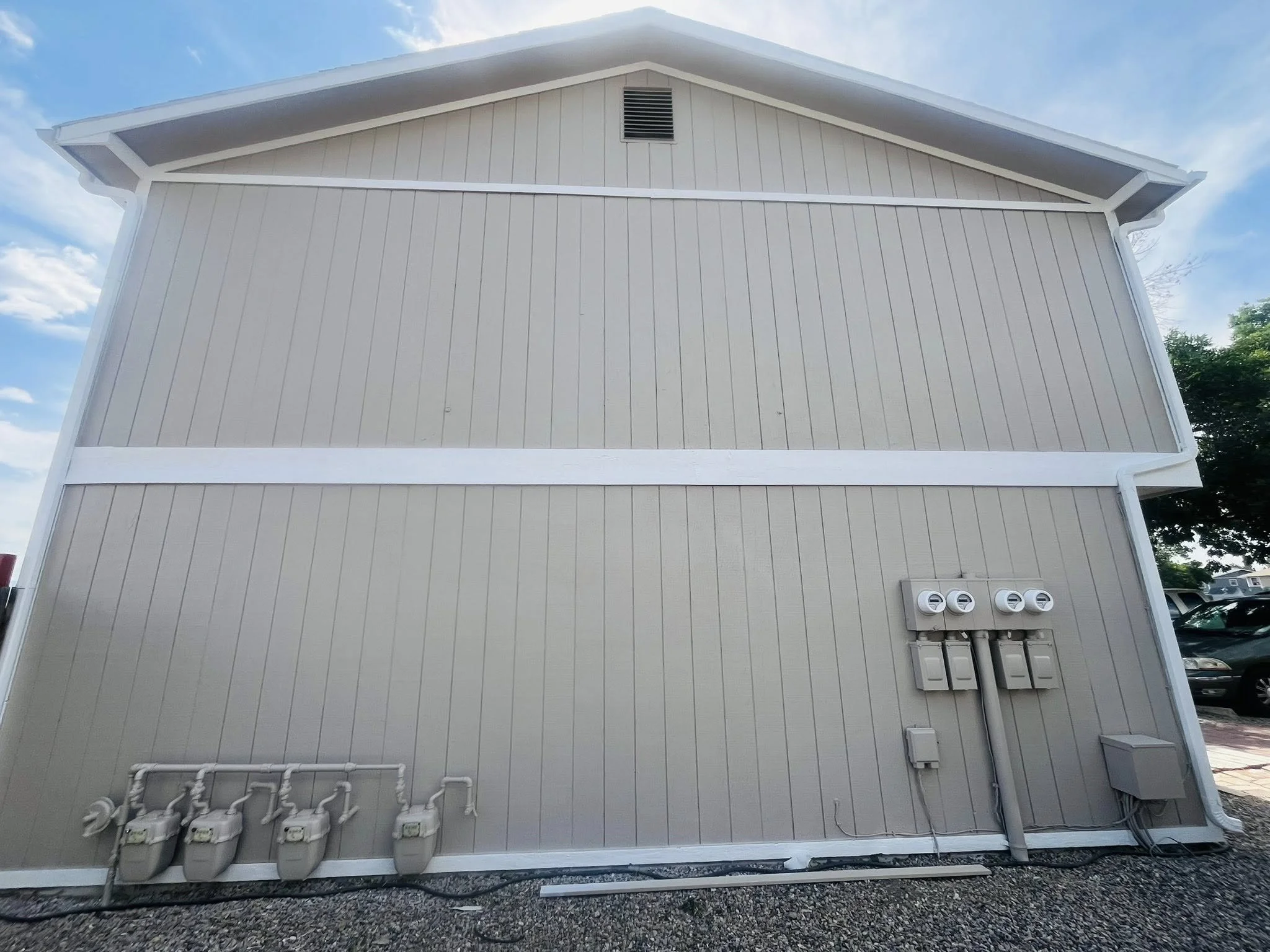 Rear view of a beige building with vertical siding, four gas meters on the left, four electric meters and electrical panels on the right, and a vent near the roof peak under a blue sky with scattered clouds.