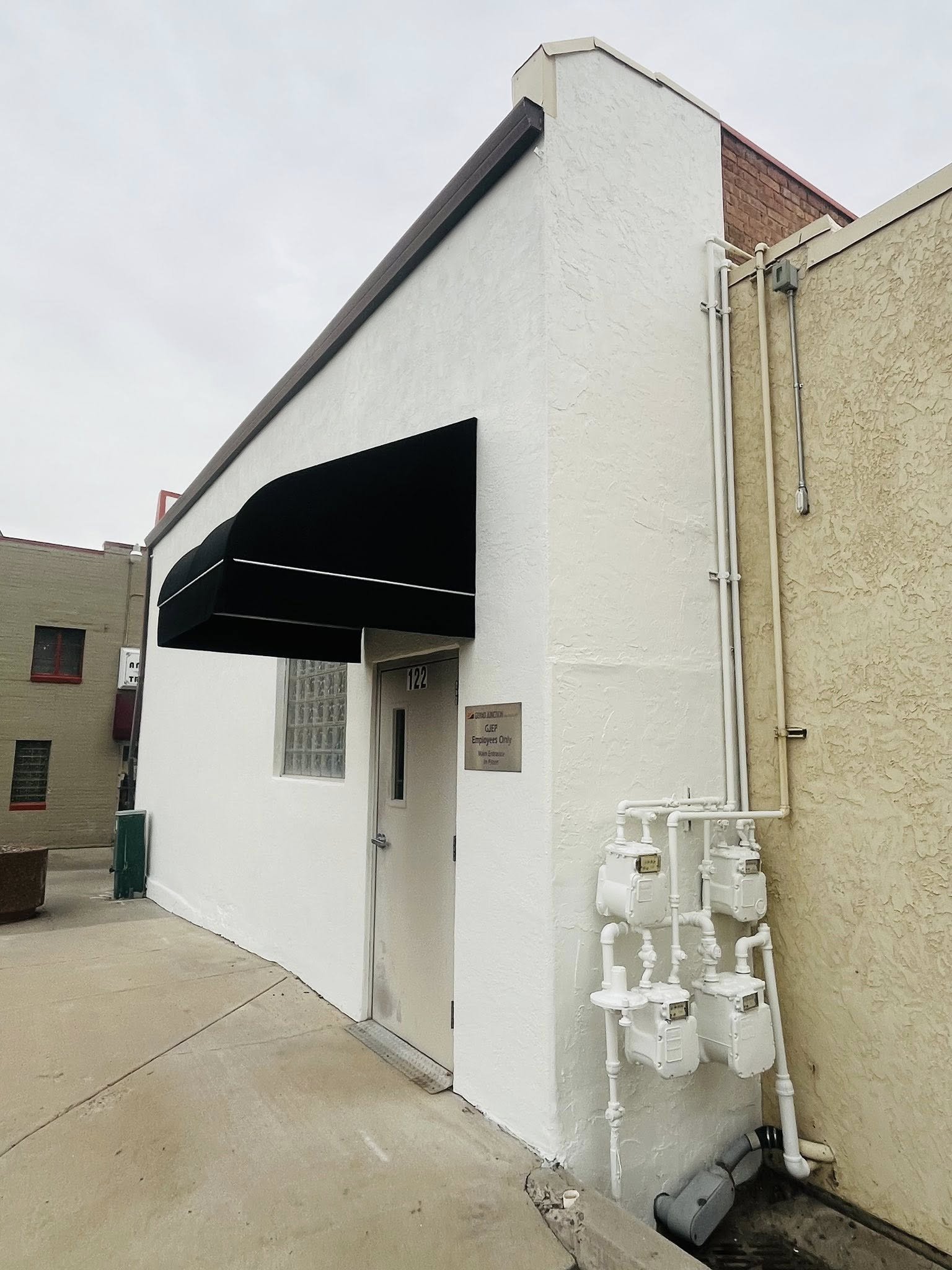 Exterior of a white building with a black awning over the door, and multiple gas meters and pipes on the wall to the right.