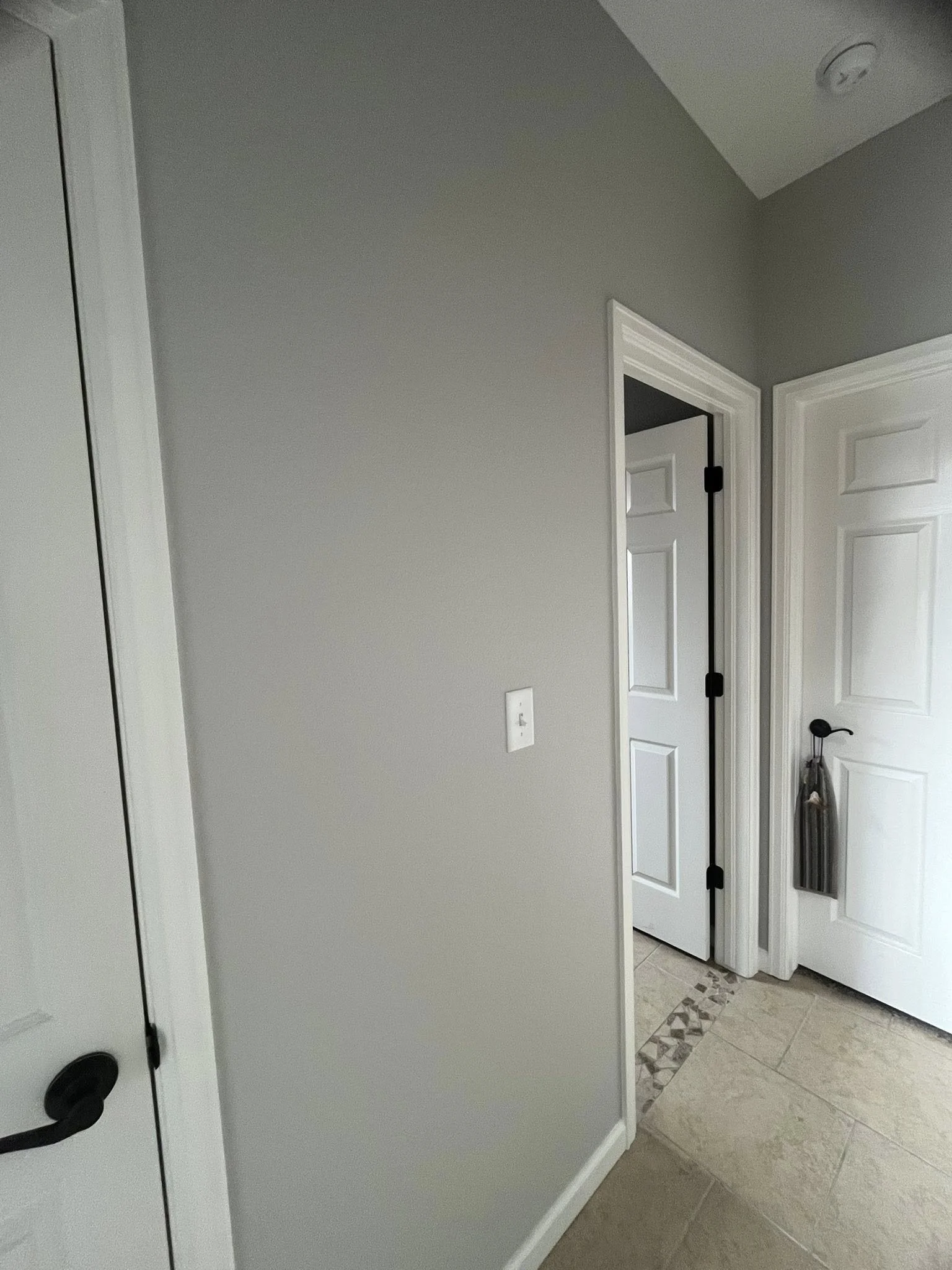 An indoor hallway with gray walls, a white door on the left, and two closed white doors on the right, with one door slightly ajar. The floor is tiled with a decorative border near the doors, and a black bag hangs on the doorknob of the door on the ri