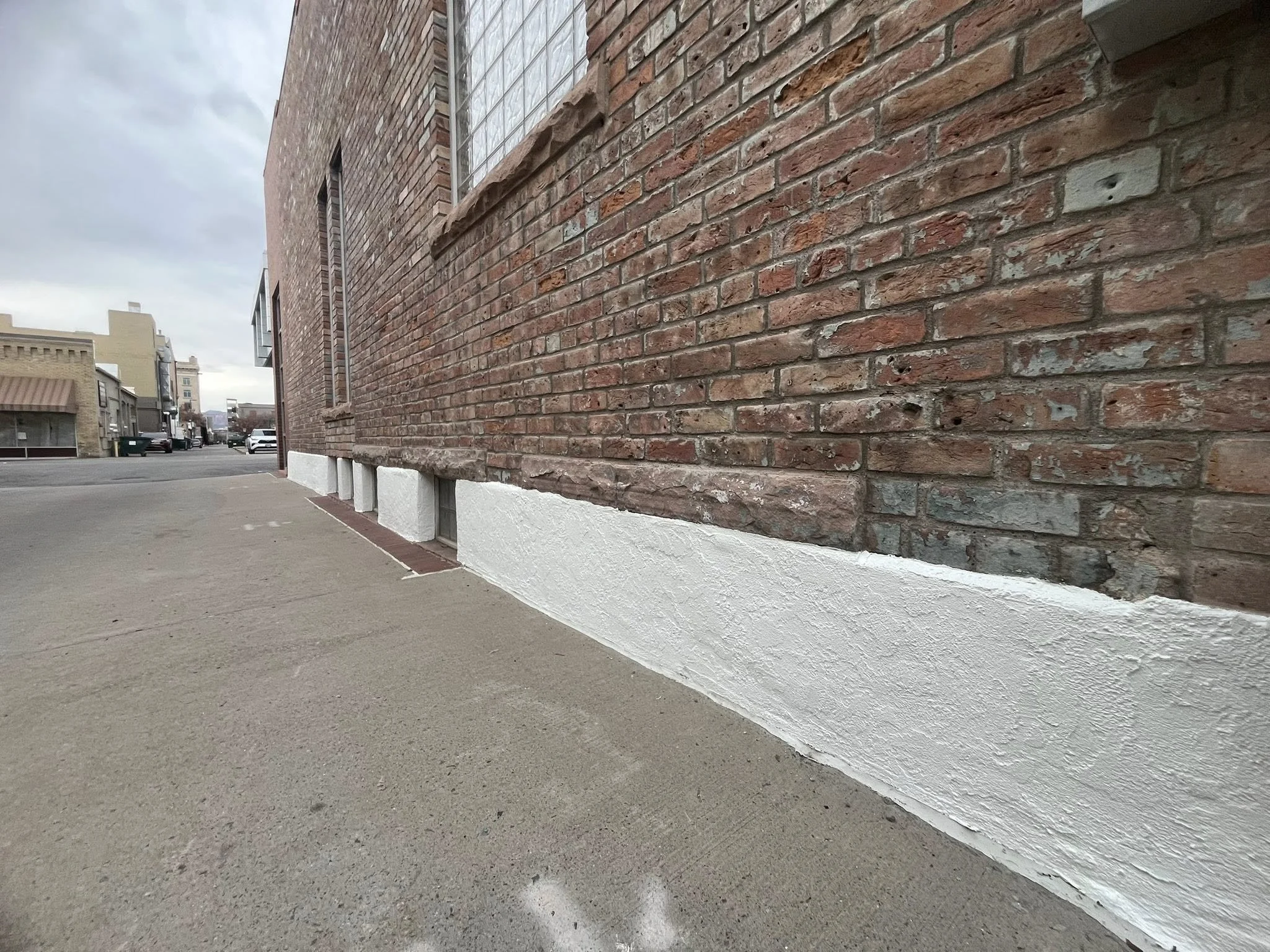 The image shows the side of a brick building with a white painted foundation. The sidewalk runs along the building, under windows, with an overcast sky in the background.