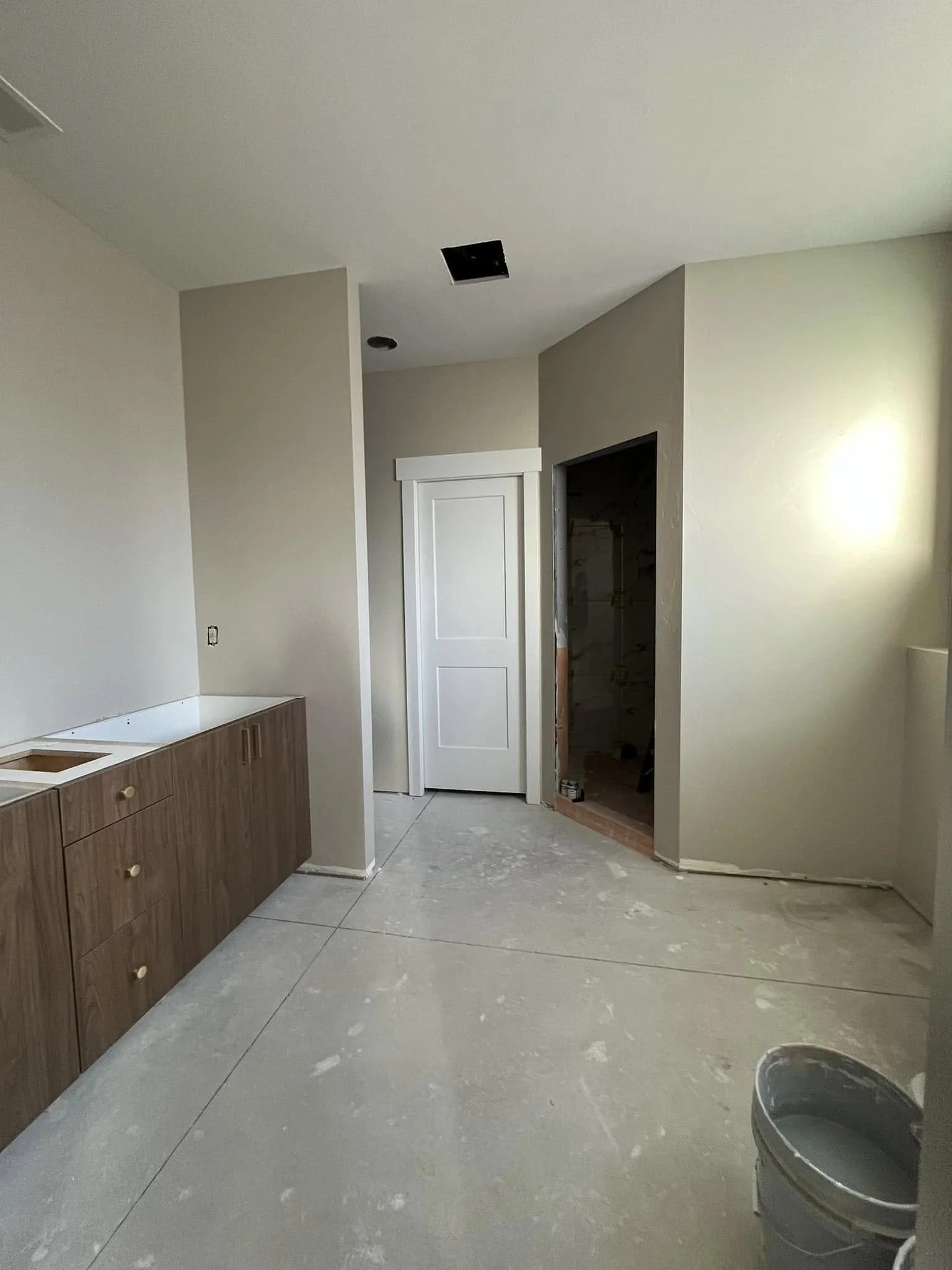 Interior of a room under construction with unfinished flooring, a wooden cabinet, a closed white door, and an open doorway leading to another space.
