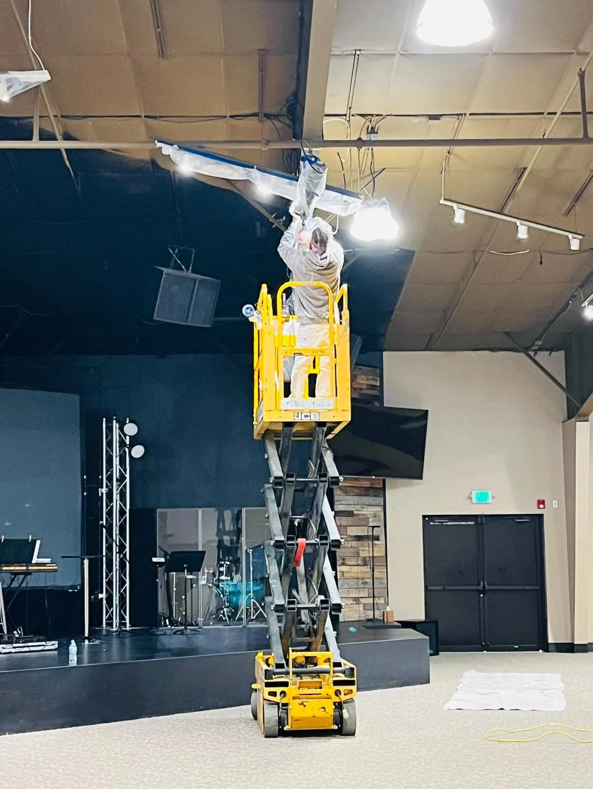 A worker standing on a yellow scissor lift, working on ceiling lighting in an indoor venue with a stage and musical instruments.