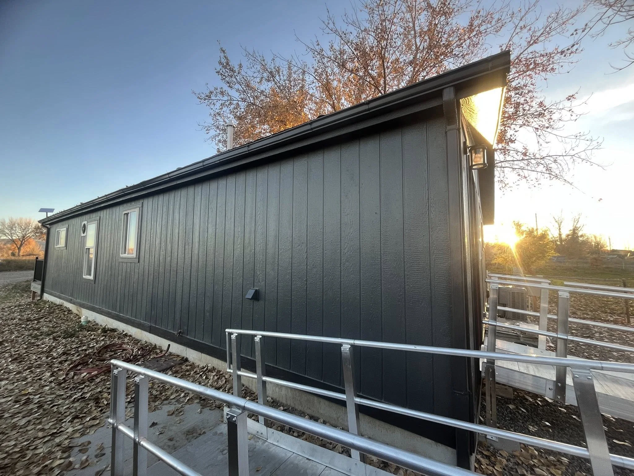 Side view of a modern tiny house with black painted wood exterior at sunset, with a metal ramp leading up to the entrance, surrounded by fallen leaves and trees in the background.