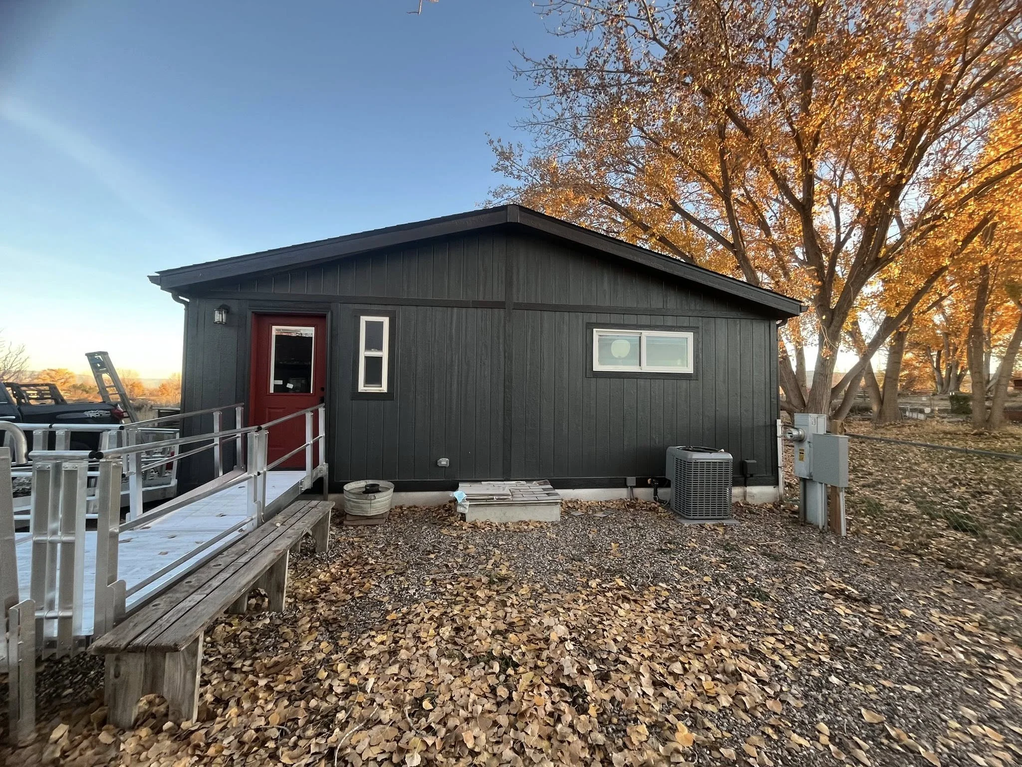 Black tiny house with a red door, small windows, a wooden ramp and a bench outside, surrounded by autumn leaves and trees with orange foliage, in late afternoon or early evening.