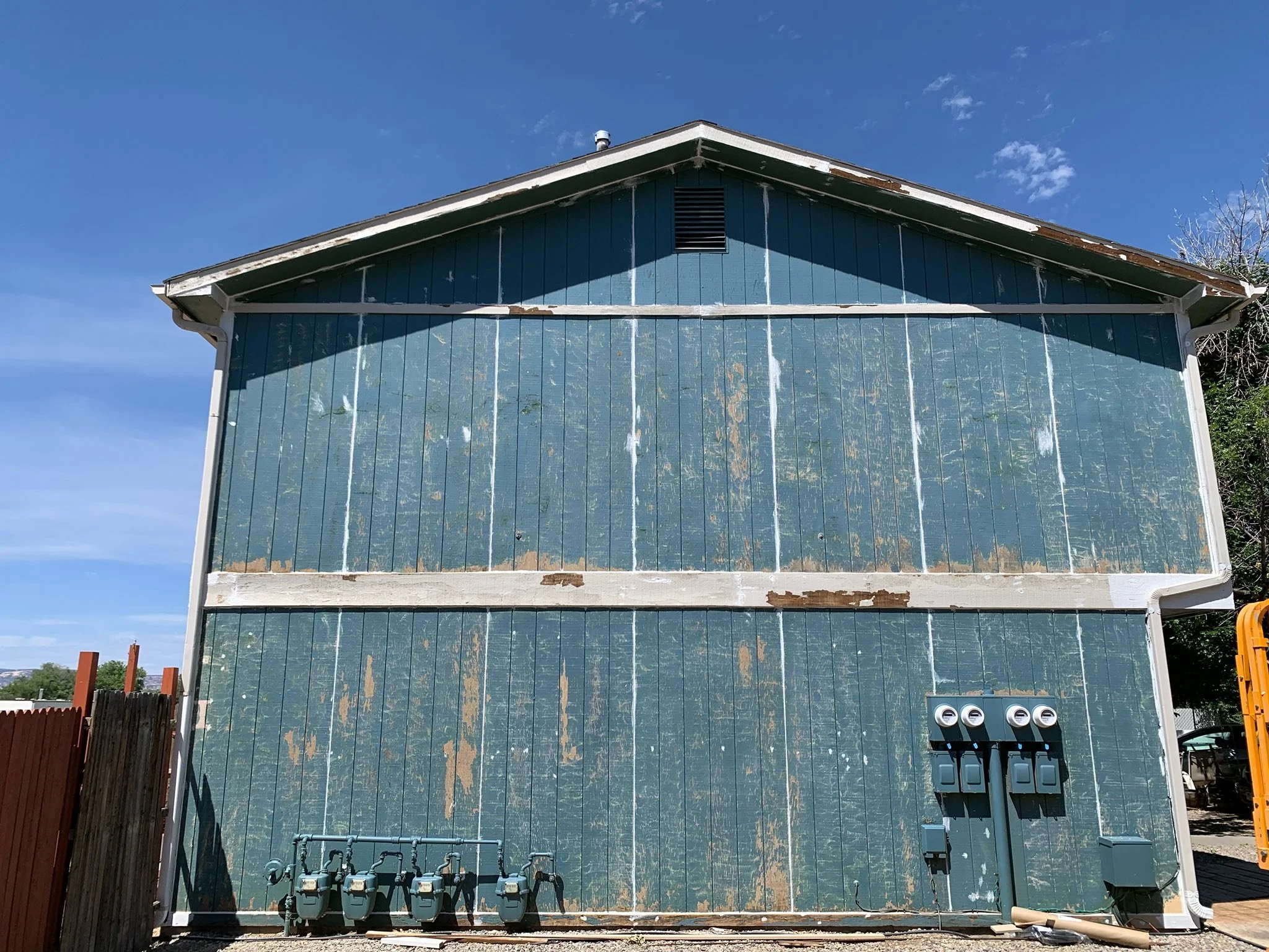 Back of a weathered blue wooden building with exposed paint and utility meters at the bottom right.