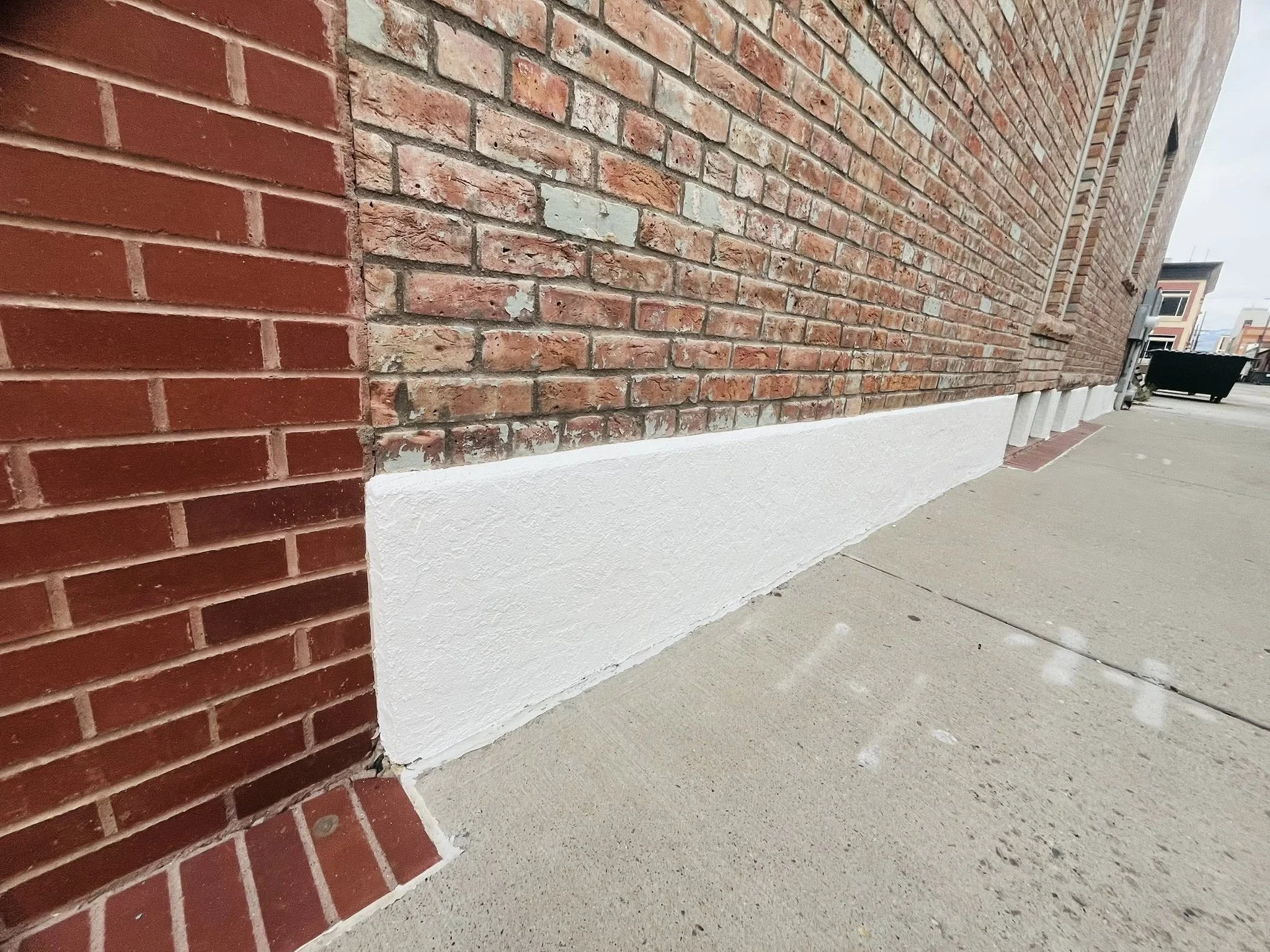 Close-up of the bottom corner of a brick building showing the contrast between the red brick and a white painted base, with a sidewalk in the foreground.