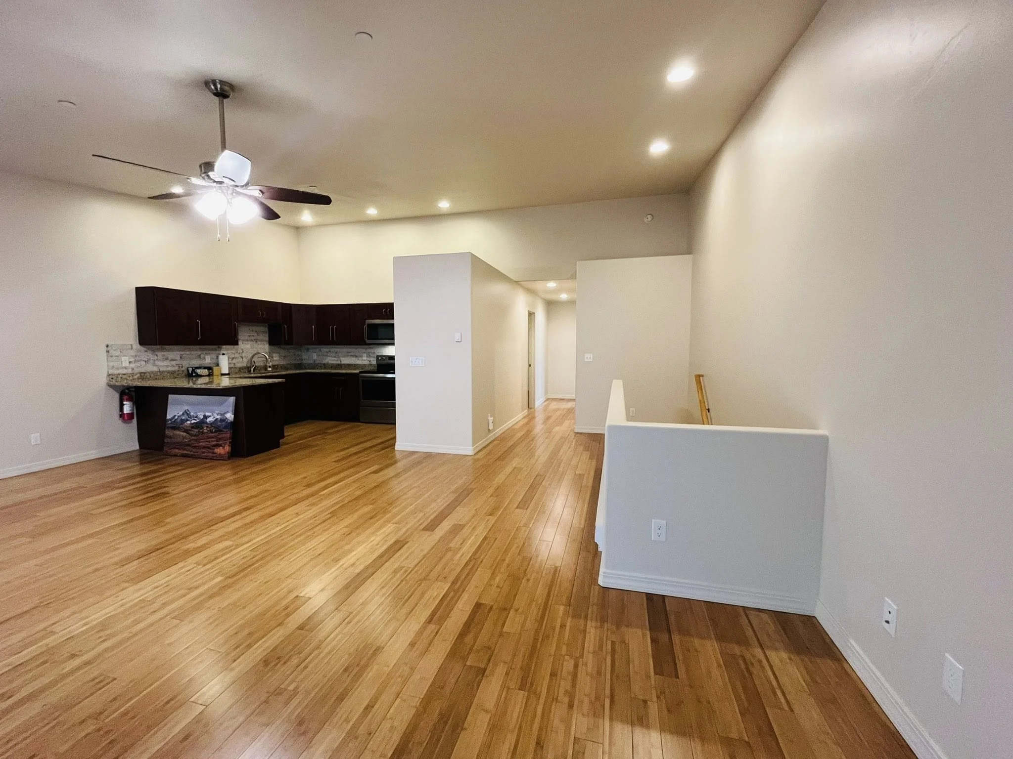 Empty living space with hardwood floors, off-white walls, kitchen in background with dark cabinets, stainless steel appliances, and a patterned backsplash, ceiling fan with lights, and a hallway leading to other rooms.
