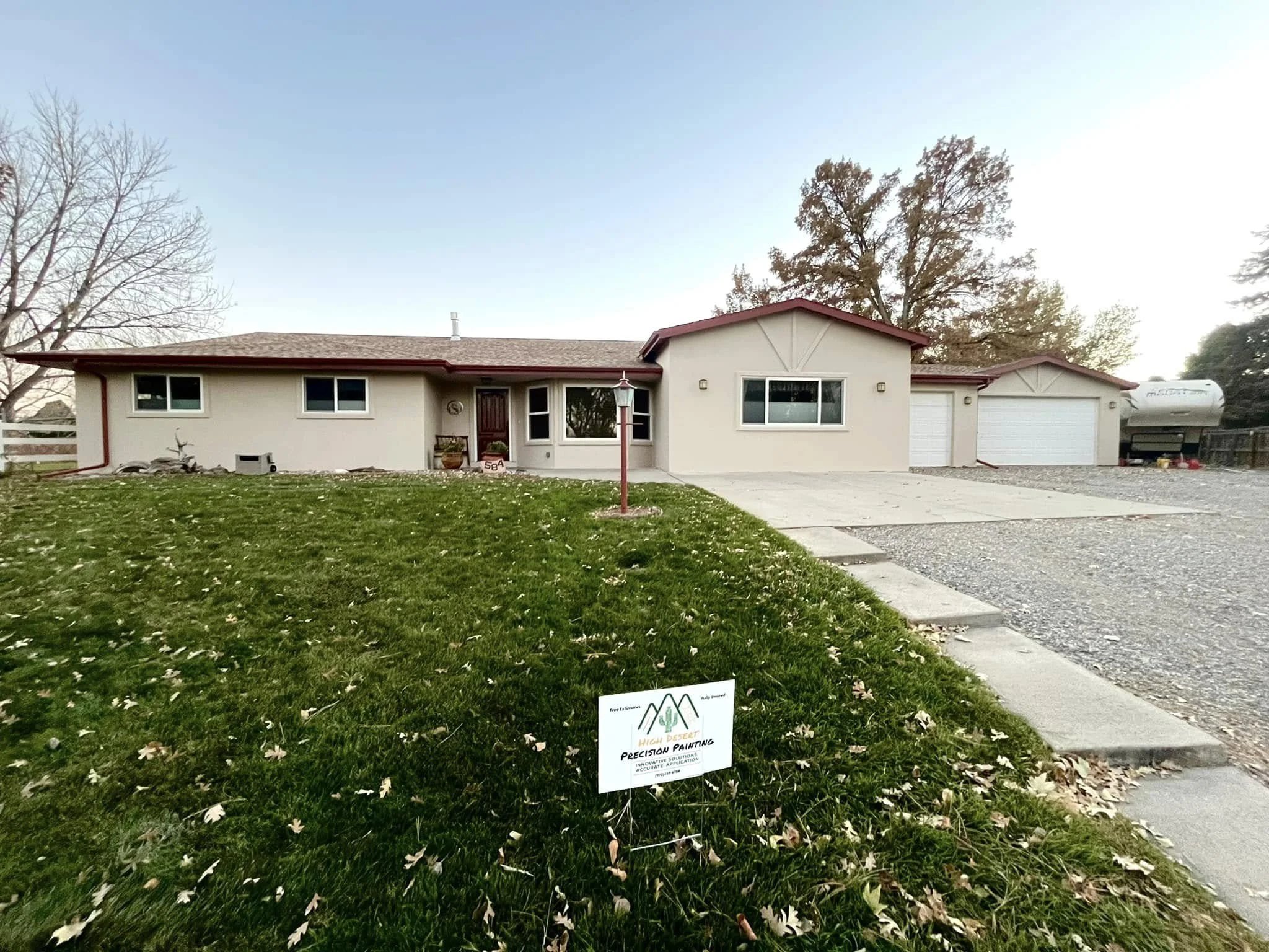 Single-story house with attached garage, front yard with green grass and fallen leaves, sign for painting company, trees in background, and clear sky.