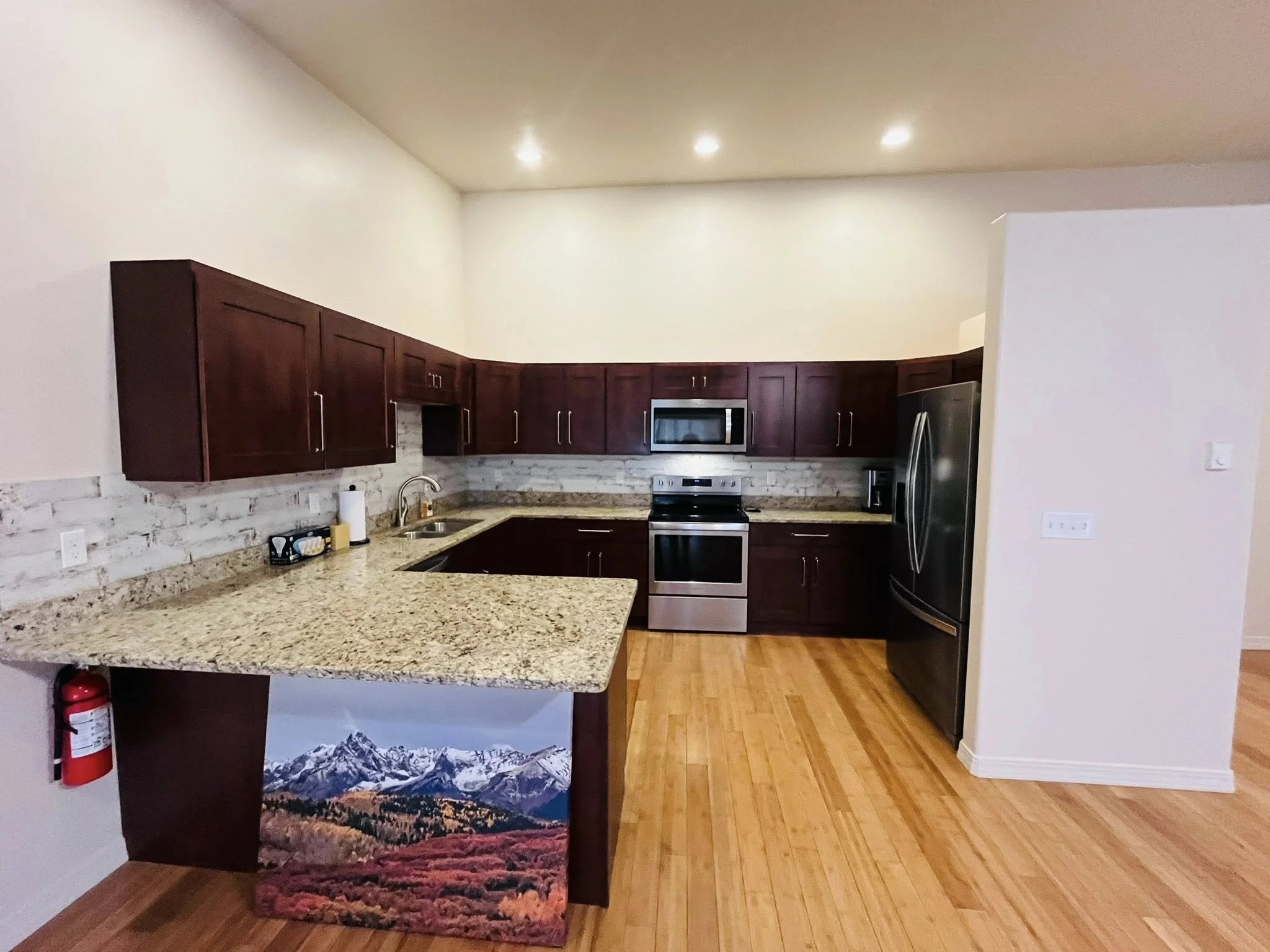 Modern kitchen with dark wood cabinets, granite countertops, stainless steel appliances, light hardwood floors, and a mountain landscape painting on a support beam.