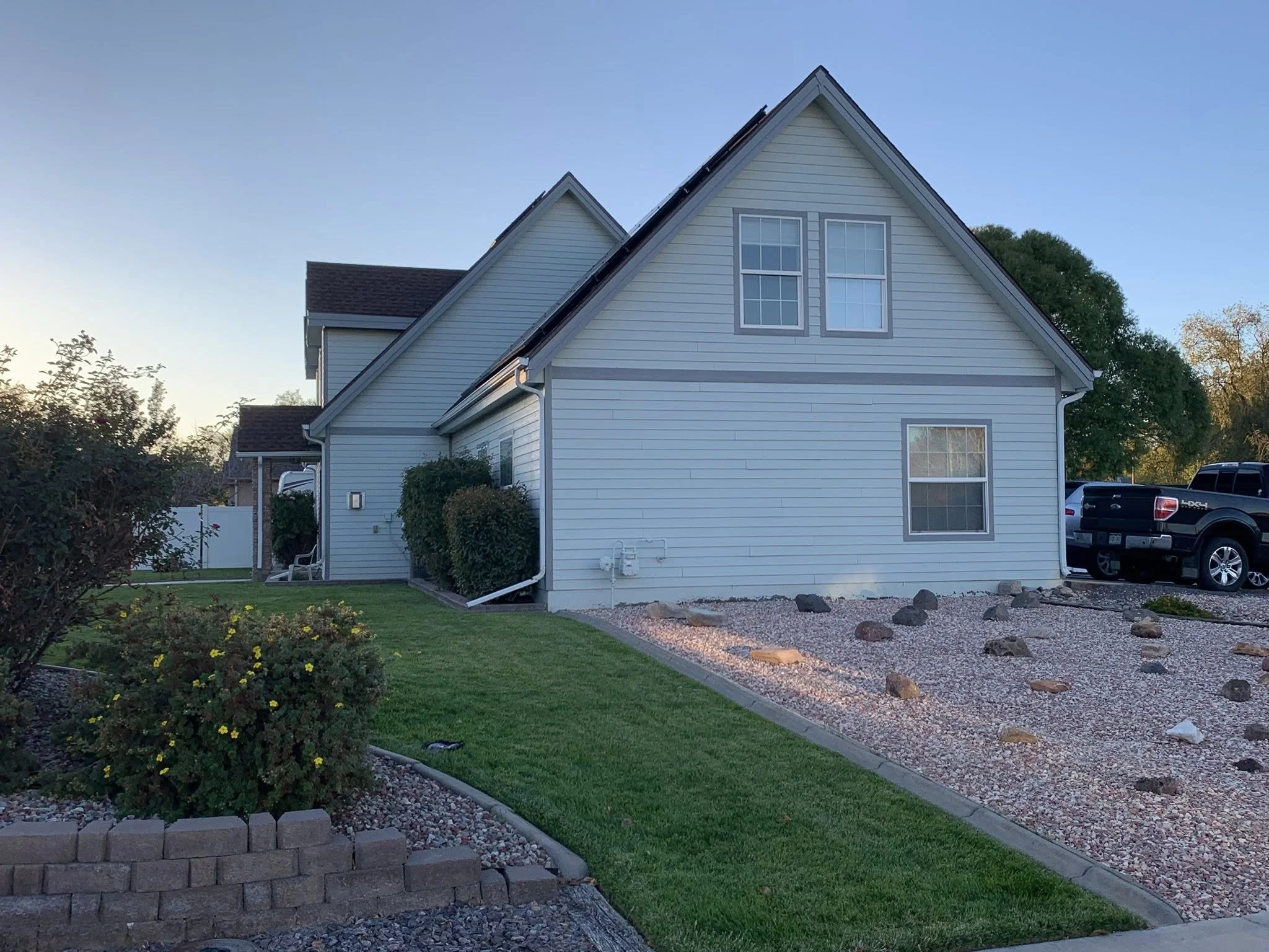 Side view of a white house with a dark roof, two large windows on the second floor, a smaller window on the first floor, and an adjacent sidewalk with a small garden containing bushes and flowers. A truck is parked on the driveway to the right.