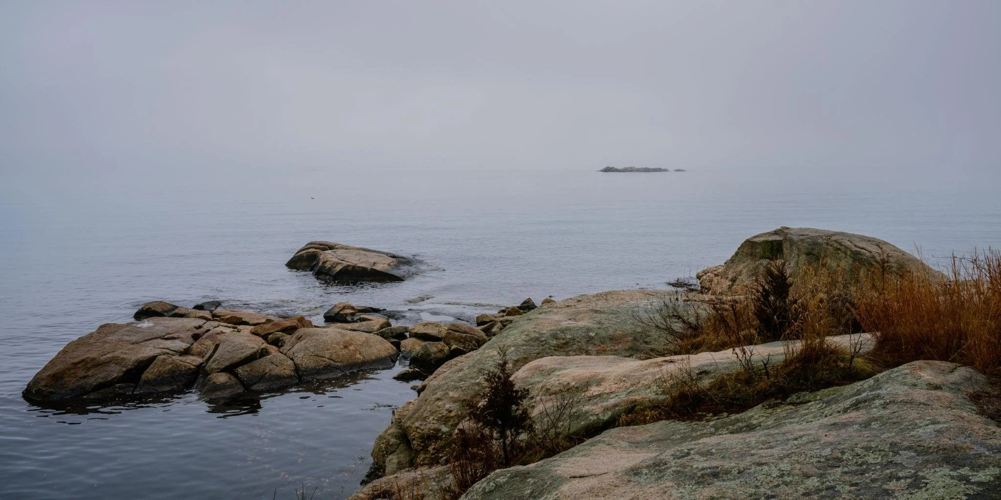 Calm lake with rocks along the shoreline, some vegetation, foggy and overcast sky, distant rocks visible in the water.