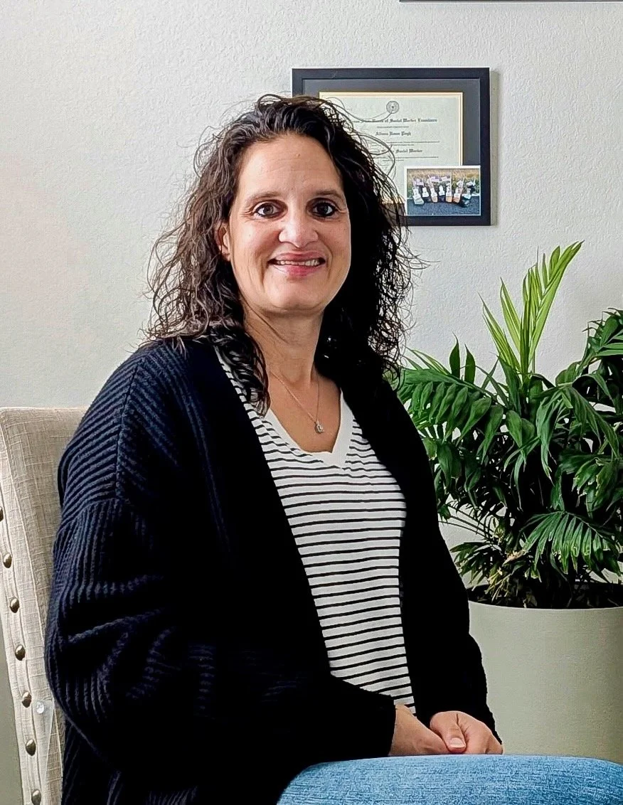 A woman with curly dark hair wearing a black cardigan and a black-and-white striped shirt, sitting in an office with framed certificates on the wall and a large green potted plant nearby.