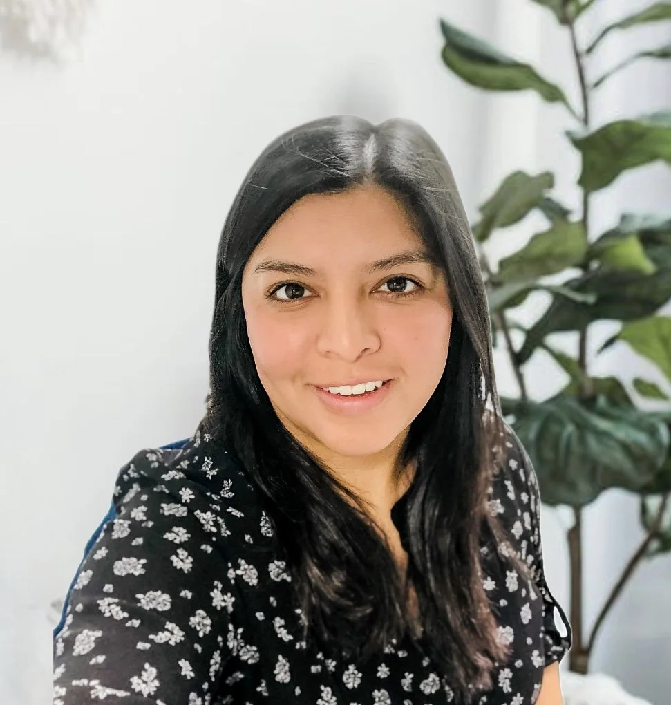 A woman with long black hair smiling at the camera, wearing a black blouse with white floral pattern, with a large leafy plant in the background.
