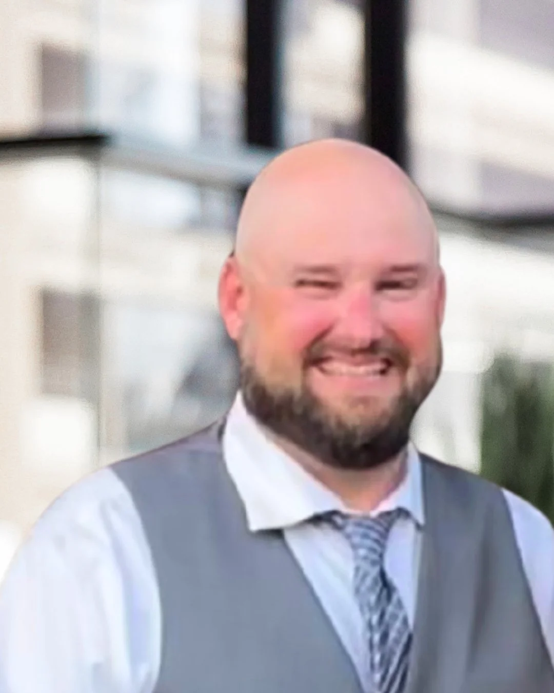 A smiling man with a bald head and beard, wearing a white shirt, gray vest, and patterned tie, standing outdoors with a building in the background.