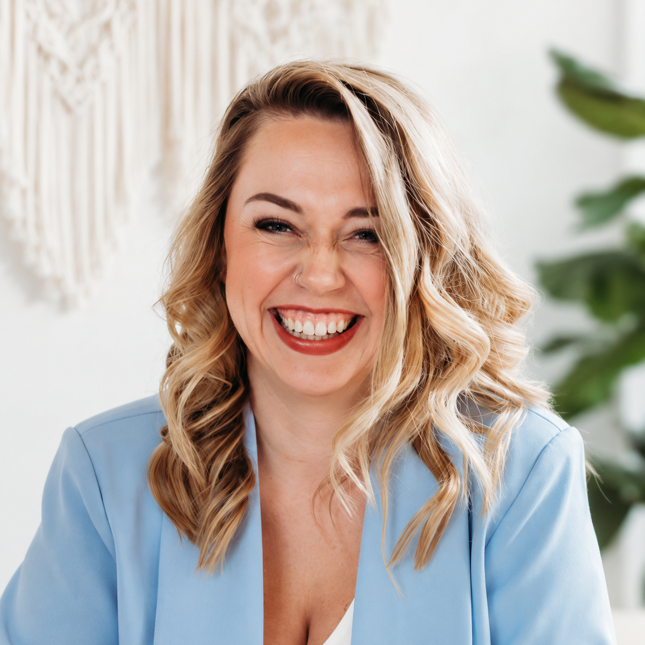 A smiling woman with wavy blonde hair, wearing a light blue blazer and jeans, standing indoors near a window with blinds, with green plants in the background.