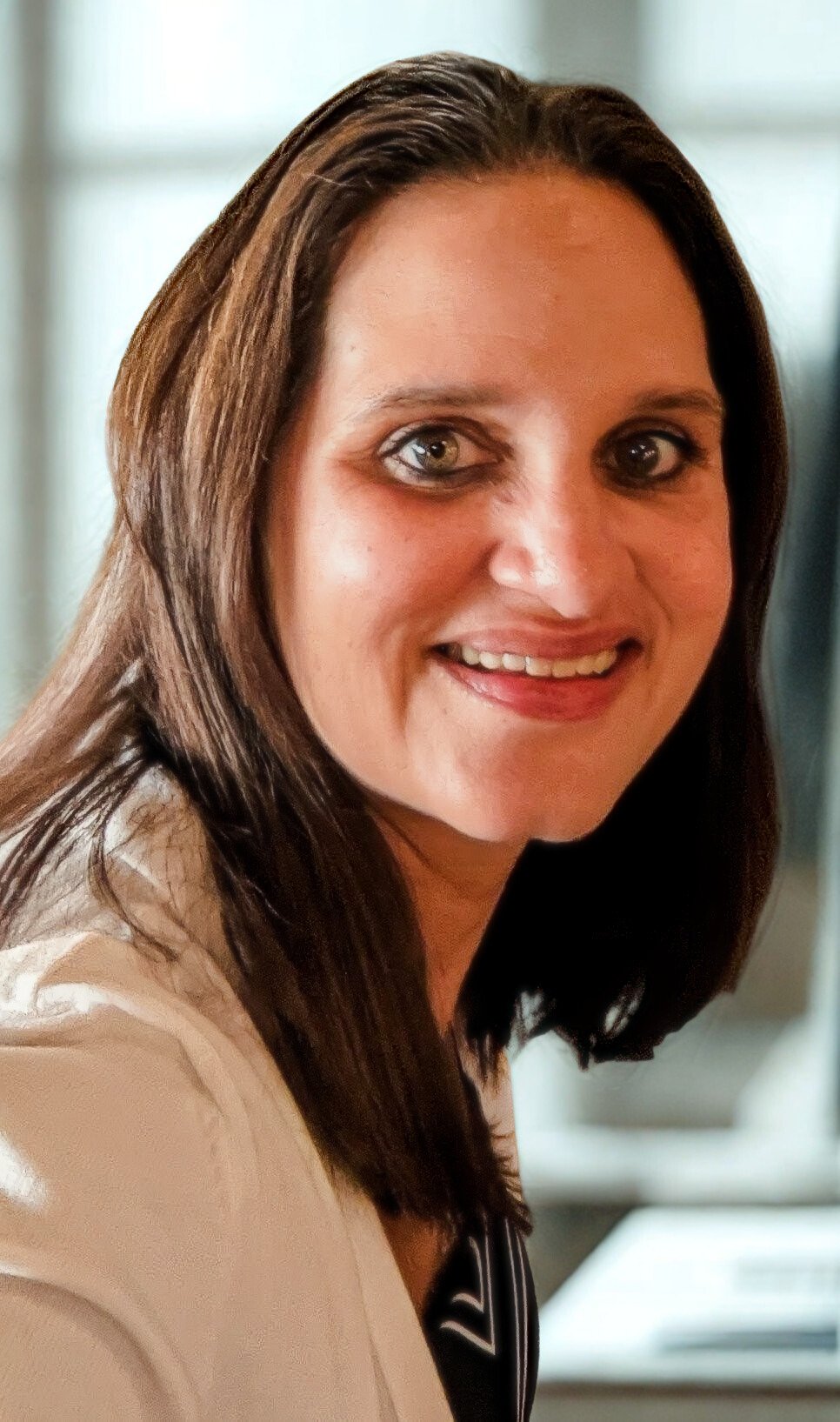 Close-up of a smiling woman with shoulder-length brown hair and light skin, indoors with a blurred background.