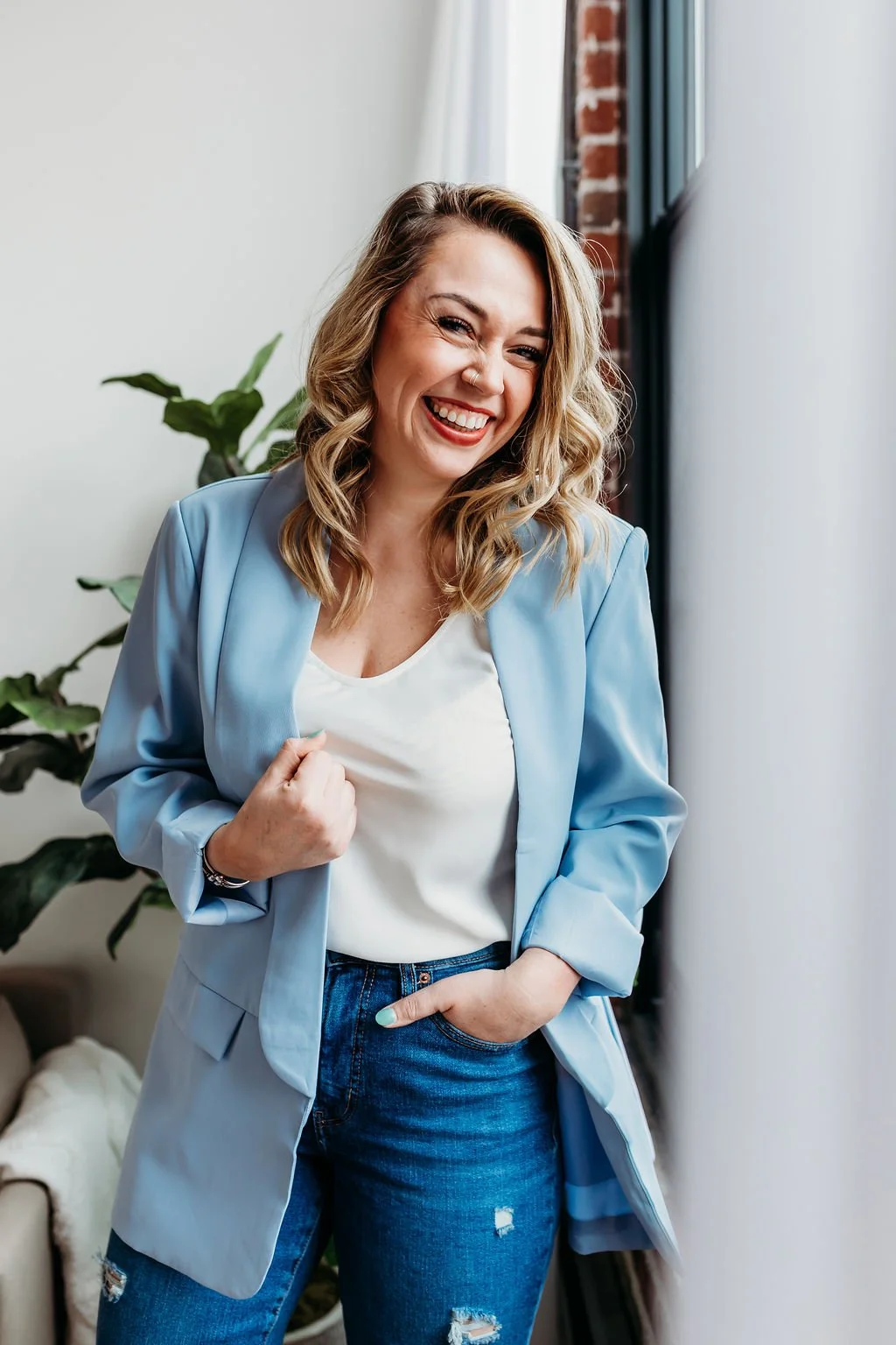 A smiling woman with wavy blonde hair, wearing a light blue blazer and jeans, standing indoors near a window with blinds, with green plants in the background.
