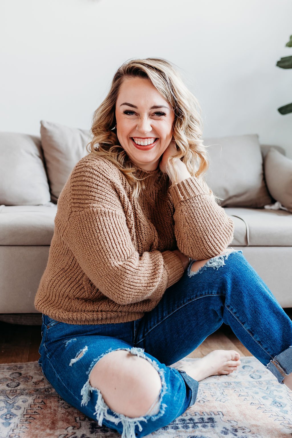 A woman with blonde curly hair smiling, sitting on the floor in front of a beige sofa, wearing a brown sweater and ripped jeans.