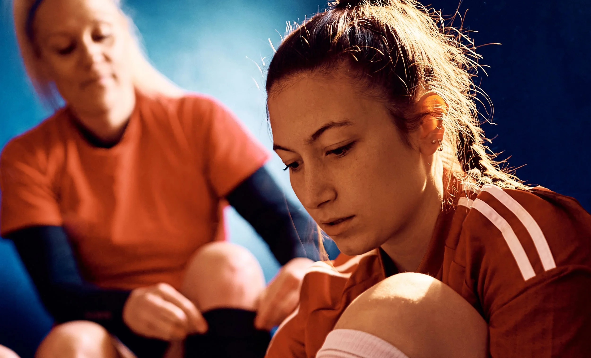 A young woman in sportswear sitting on the ground, looking down with a serious expression, with another woman in sportswear in the background.