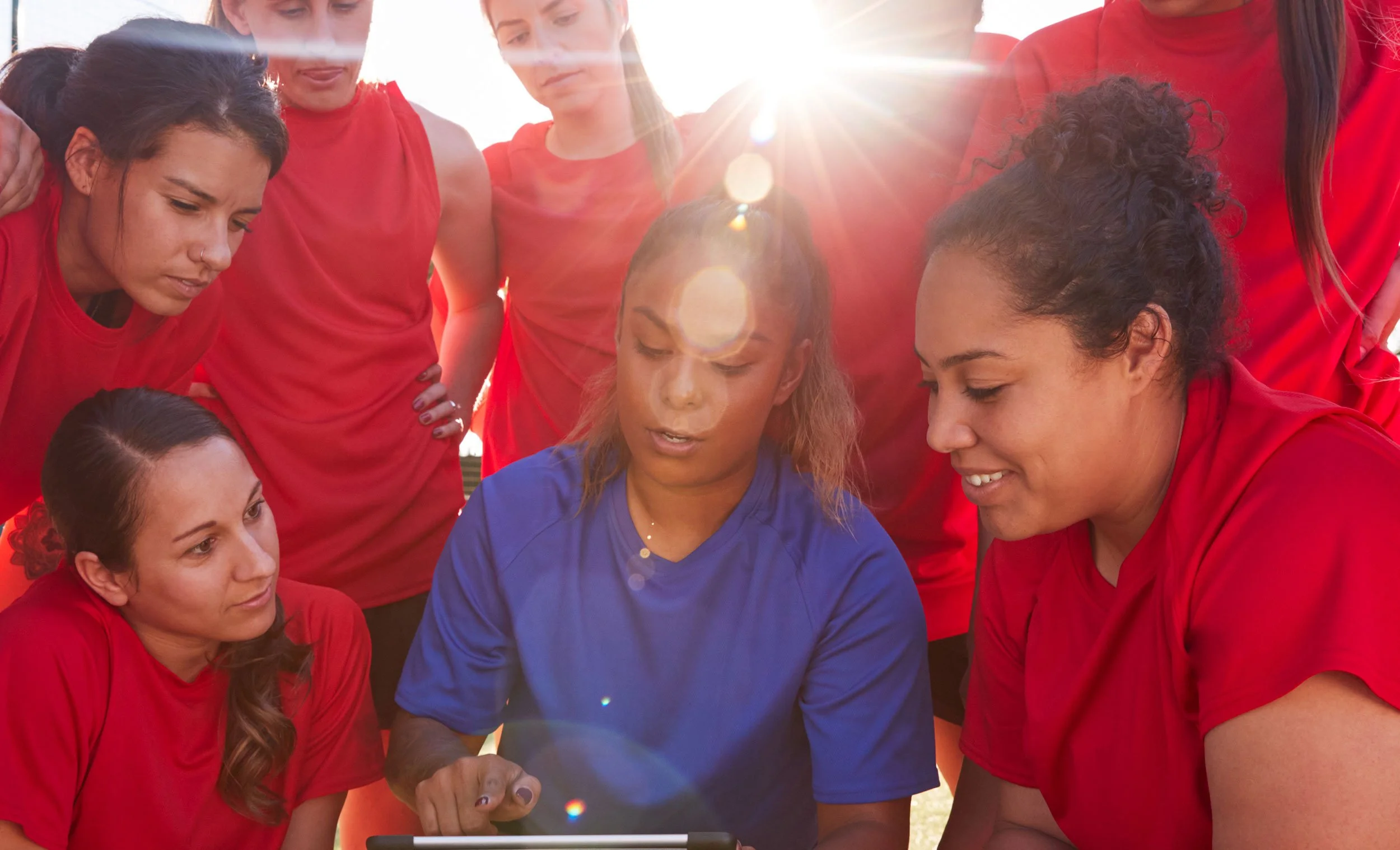 A group of female soccer players, mostly wearing red jerseys, gathered around a woman in a blue jersey, looking at a smartphone outdoors during sunset.