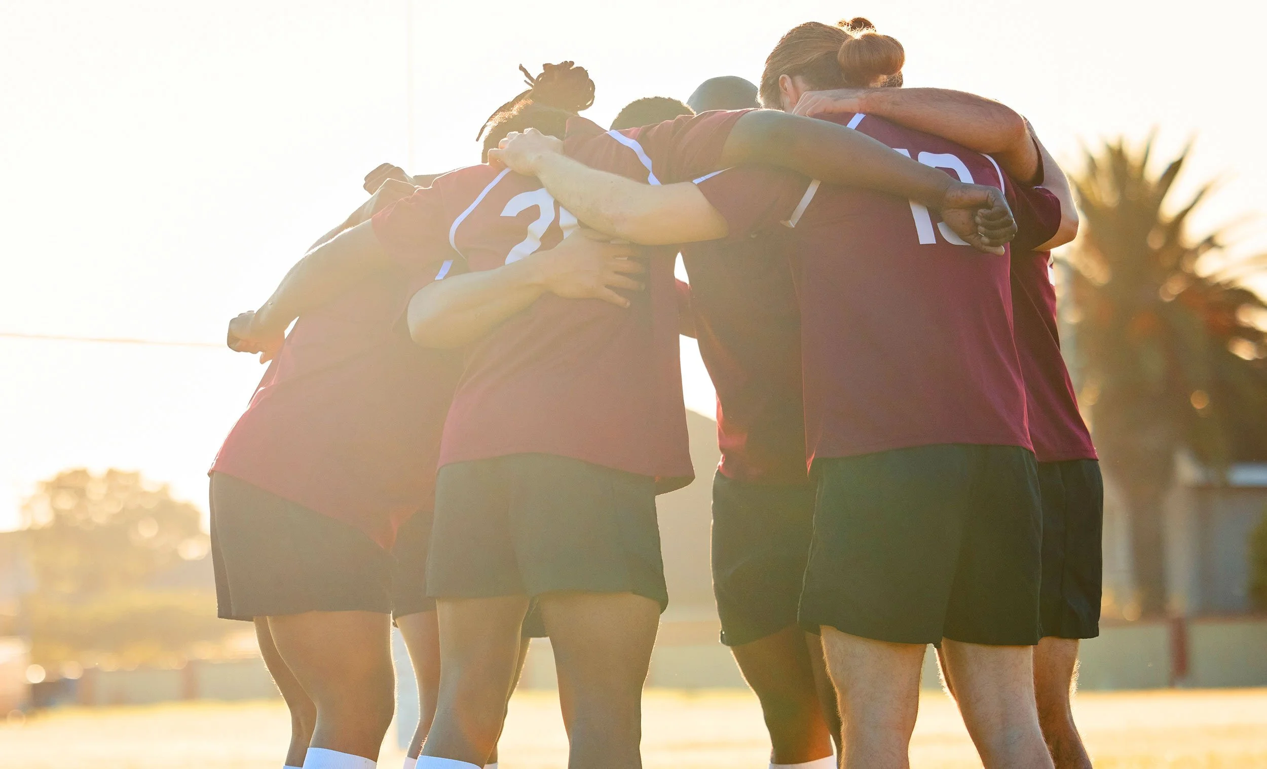 A group of rugby players huddled together in a team cheer on an outdoor field during sunset.