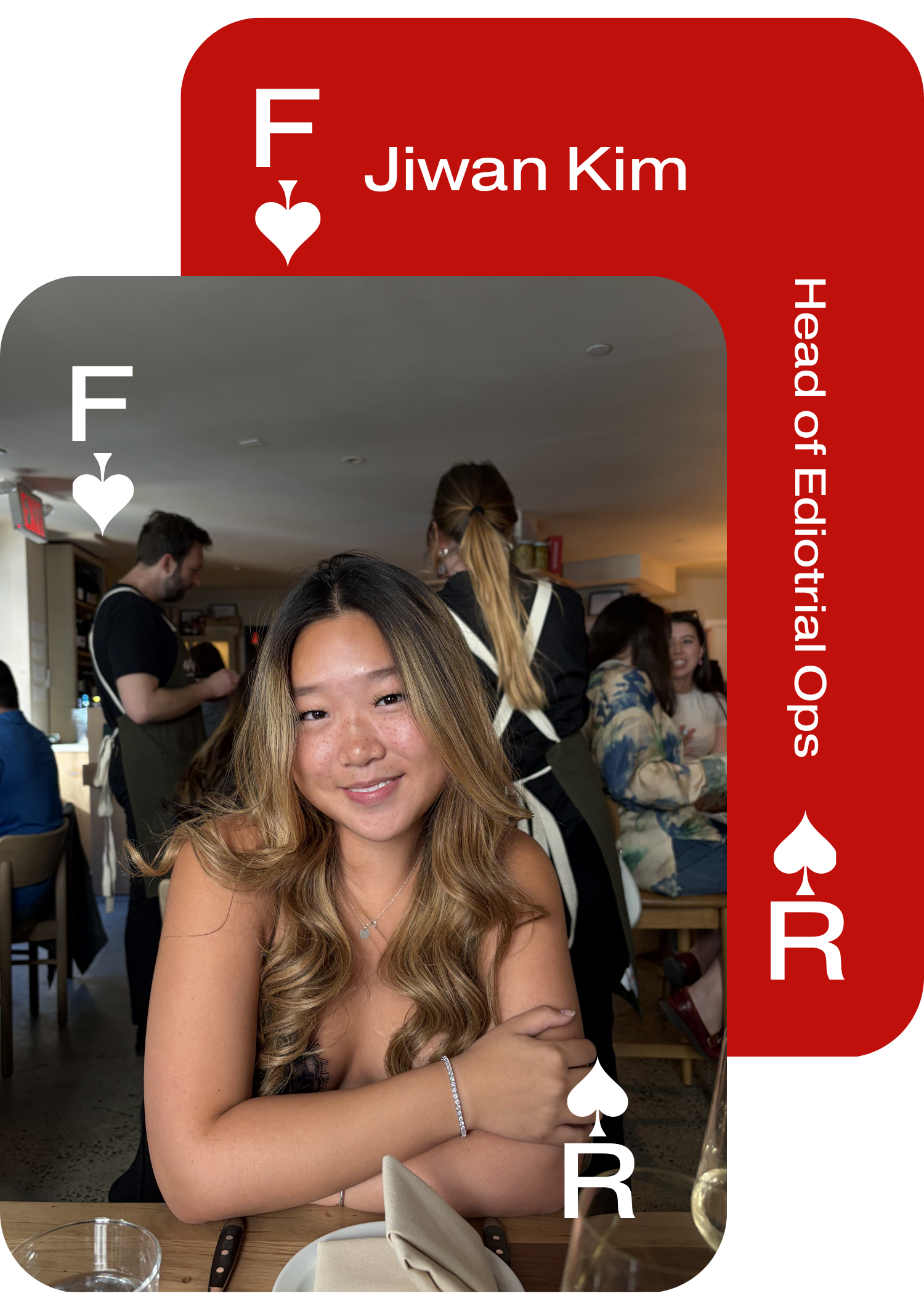 A young woman with long, wavy, blonde hair and a bright smile sitting at a table in a restaurant. She is wearing a black dress, a silver bracelet, and a necklace. There are glasses, a napkin, and a knife on the table. In the background, other diners are visible, including a man with a beard and dark hair, and women with long hair, one with her hair tied back.