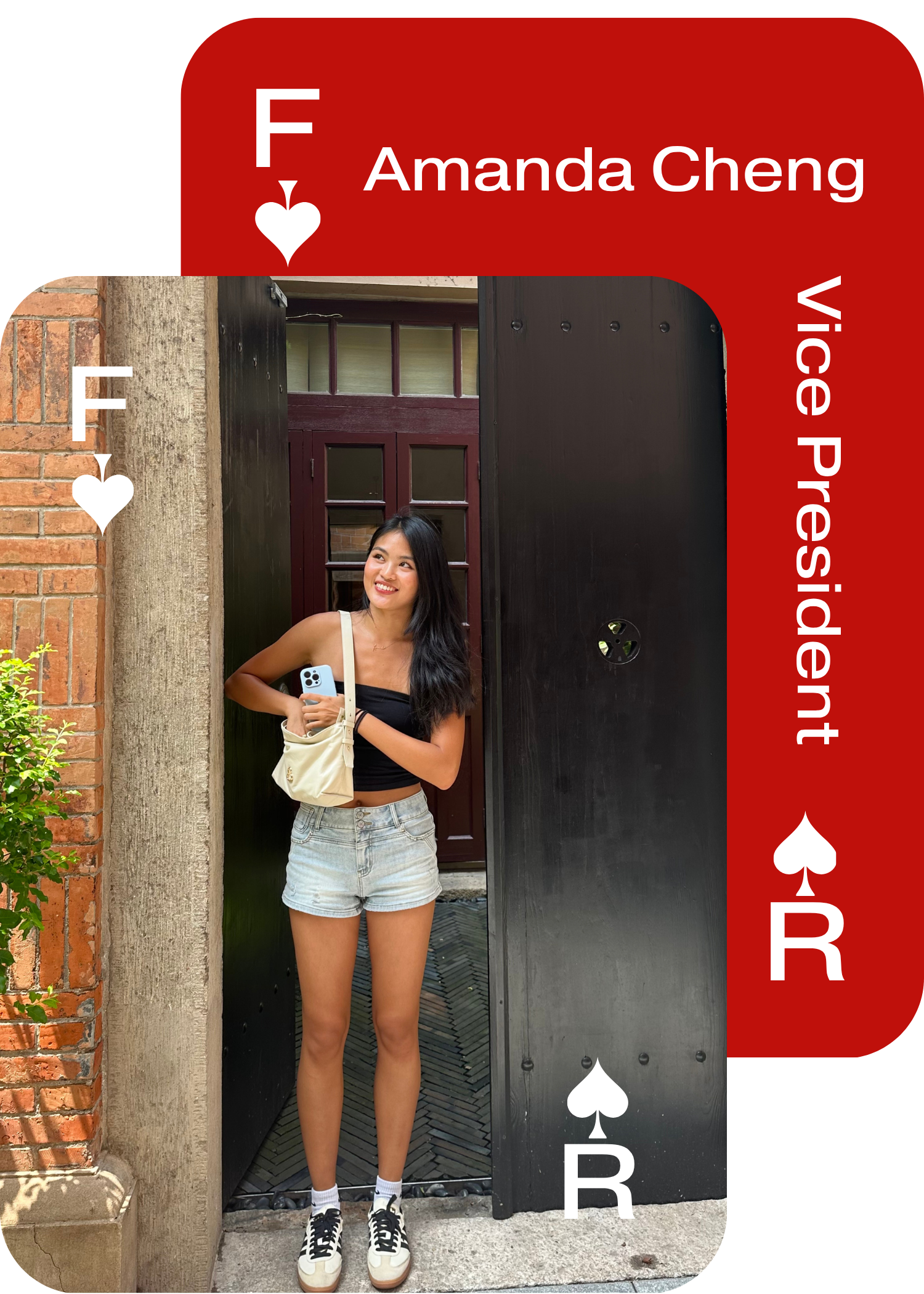 Young woman with long dark hair, wearing a black tank top, denim shorts, white socks, and black and white sneakers, standing in front of an open black door with brick and concrete walls, smiling and holding a white bag and a phone.