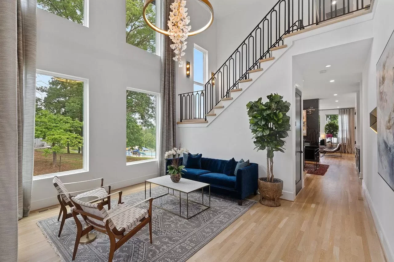 Living room with large windows, a dark blue sofa, a coffee table, wooden chairs, a patterned rug, a tall potted plant, and a staircase leading to an upper level.