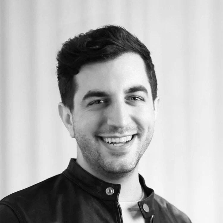 Black and white portrait of a smiling man with dark hair, wearing a dark jacket with a zipper.