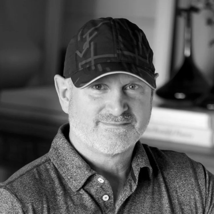 Black and white photo of a middle-aged man wearing a cap and a collared shirt, smiling softly, with a bookshelf and a table lamp in the background.