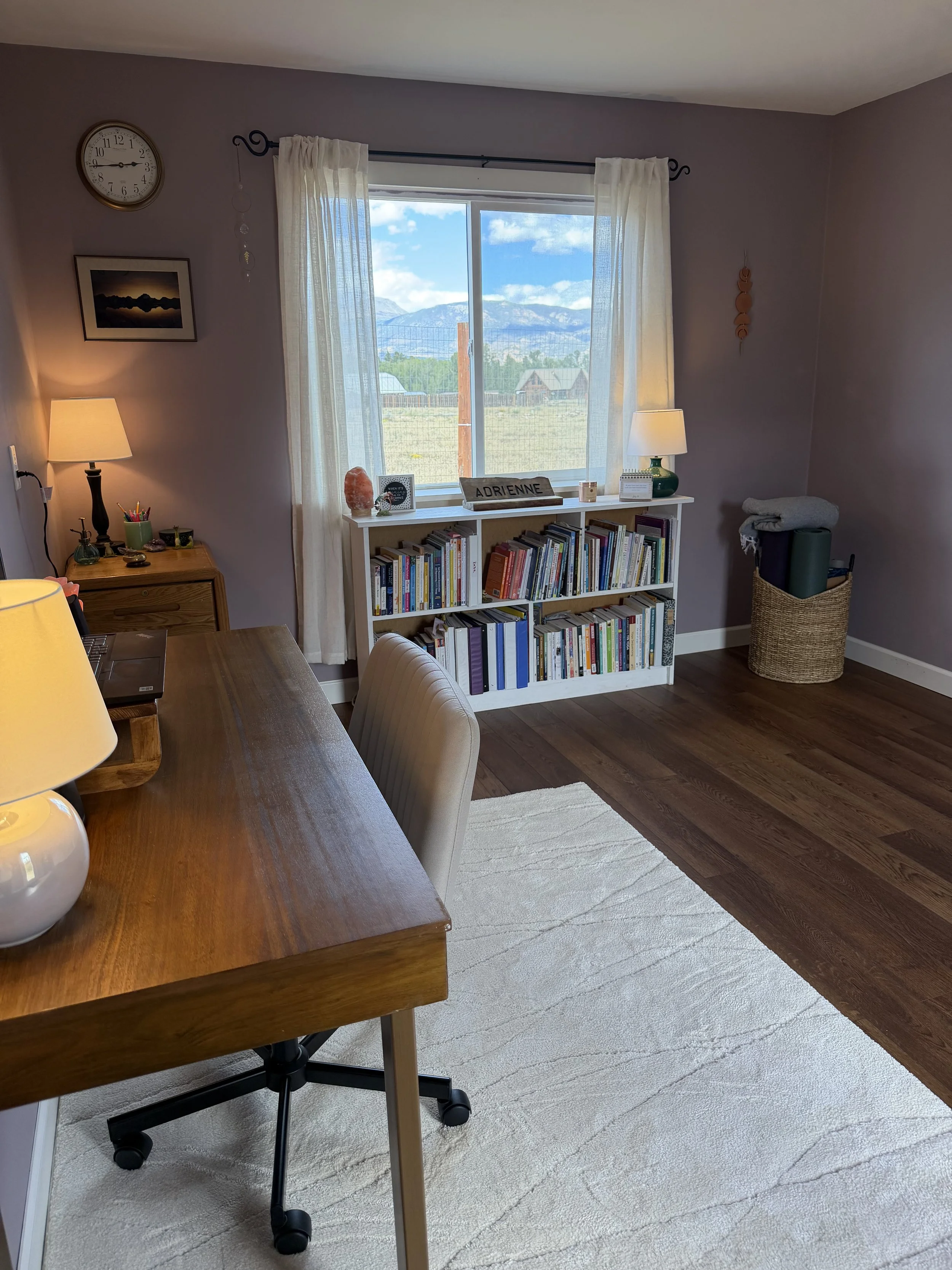 Warm telehealth workspace with a wooden desk, bookshelf, soft lighting, and a large window.