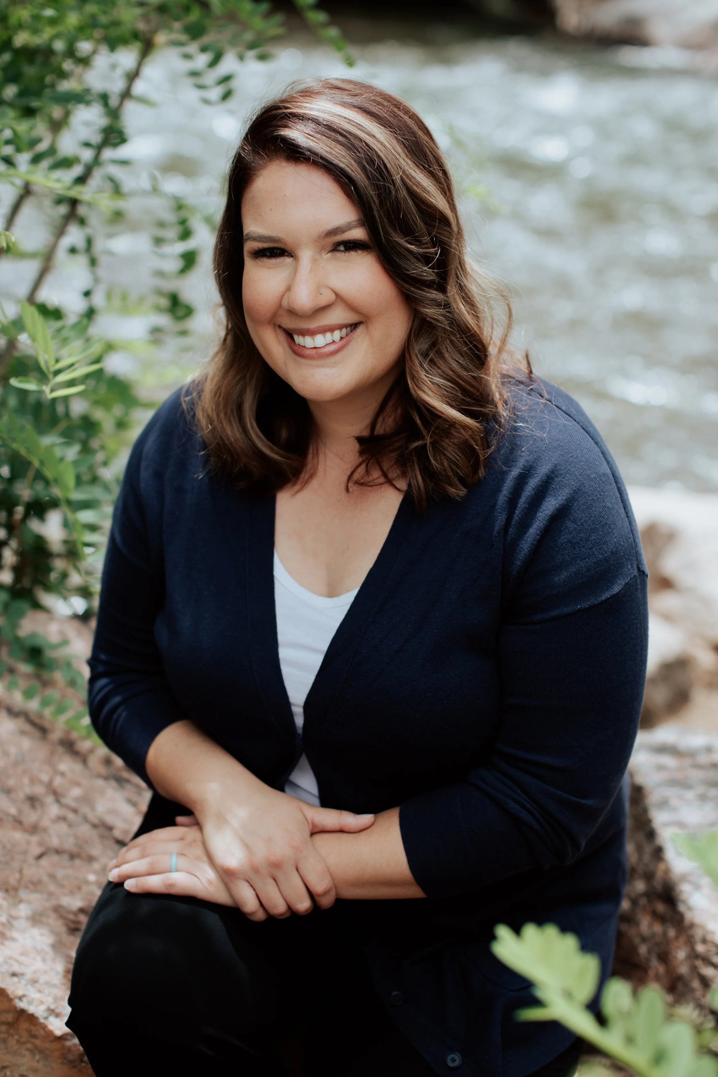 A woman with shoulder-length brown hair, smiling, sitting outdoors near a body of water, wearing a navy blue cardigan over a white top.