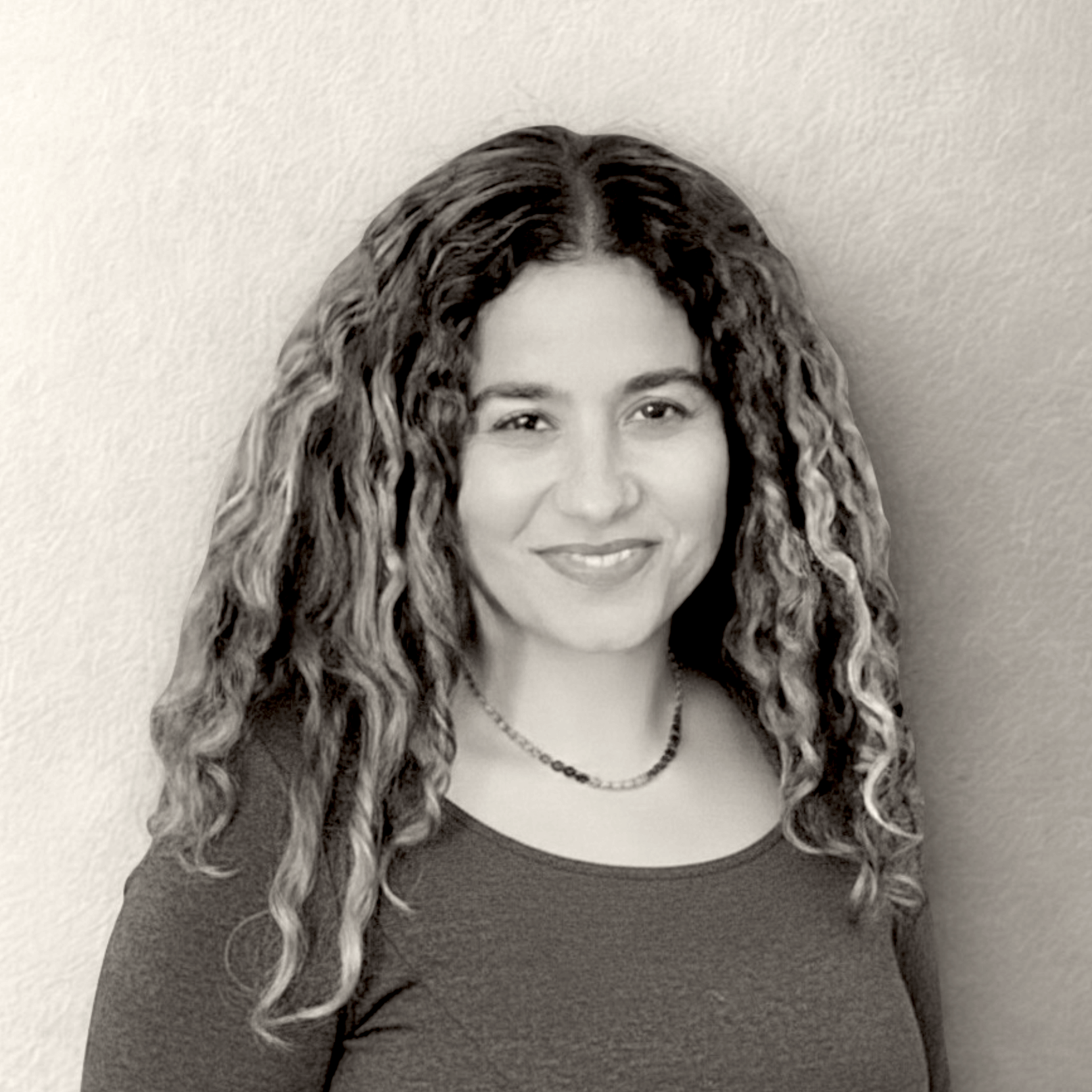 Black and white photo of a woman with curly hair, wearing a necklace and a dark top, smiling at the camera, against a plain wall background.